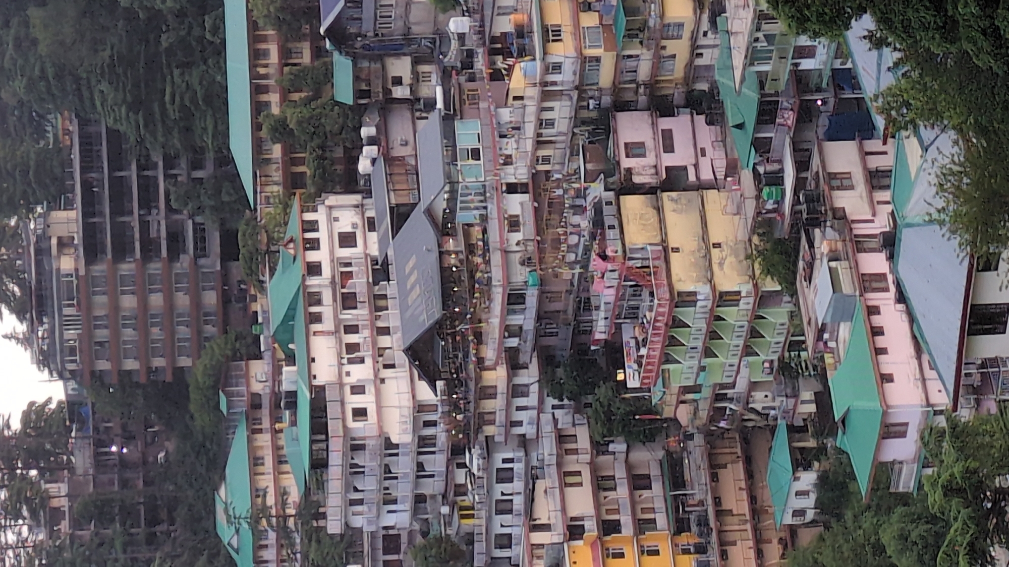 The image captures a dense urban landscape, characteristic of a hill town, seen from an elevated perspective. The setting is a valley or hillside densely packed with multi-story buildings, interspersed with lush green trees, primarily conifers. The location context of Dharamshala, India, is consistent with the visual cues.
The scene is dominated by a multitude of structures varying in size, age, and color. Many buildings feature vibrant facades in shades of yellow, pink, beige, white, and light blue. A significant number of roofs are distinctively colored in green or light blue, with some being grey or flat. In the far left (if rotated to a normal orientation), a large, multi-story concrete structure appears to be either under construction or unfinished, with exposed beams and scaffolding-like elements.
Scattered throughout the mid-ground, numerous balconies and windows are visible. Clothes lines with drying laundry can be seen on some balconies, indicating residential use. Notably, strings of colorful prayer flags, a common sight in Himalayan regions, are strung between several buildings in the central part of the image, adding a cultural identifier. A few red-colored external staircases or ladders are also visible on some buildings.
On one prominent grey rooftop in the mid-left area, white text is clearly discernible, spelling out "HOTEL VILLAGE VIEW" or "HOTEL VILLAGE INN".
The lighting is soft and diffused, suggesting either an overcast day or the early morning/late afternoon hours. No harsh shadows are present, contributing to a muted overall tone. There are no identifiable people or specific events taking place that can be discerned from this distant view. The general atmosphere is that of a bustling, settled hill community.