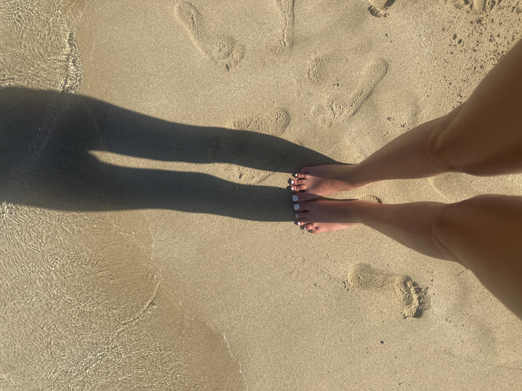 A first-person perspective shot of a person's bare feet and lower legs standing on a sandy beach. The feet are adorned with white-painted toenails and are positioned in the foreground. Behind the feet, footprints are visible in the sand, some partially submerged in shallow, clear water with a rippled surface reflecting sunlight. A long shadow of the person's legs is cast across the sand and into the water. The scene is located on a beach in Protaras, Cyprus, identifiable by the characteristic golden sand and the crystal-clear waters of the Mediterranean Sea.