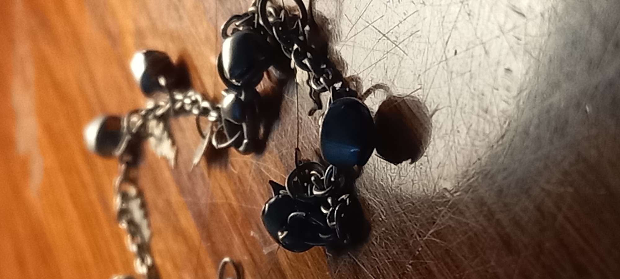 The image shows a close-up, low-angle view of a silver-toned chain bracelet with dark blue beads, resting on a wooden surface with visible scratches. The bracelet appears to be a piece of jewelry, possibly an anklet or a bracelet. The lighting is dim, creating shadows and highlighting the reflective surfaces of the metal and beads. There is no discernible connection to Kryvyi Rih, Ukraine, as the image is a close-up of an inanimate object and its setting, lacking any contextual information or landmarks that would identify its location within a specific city.