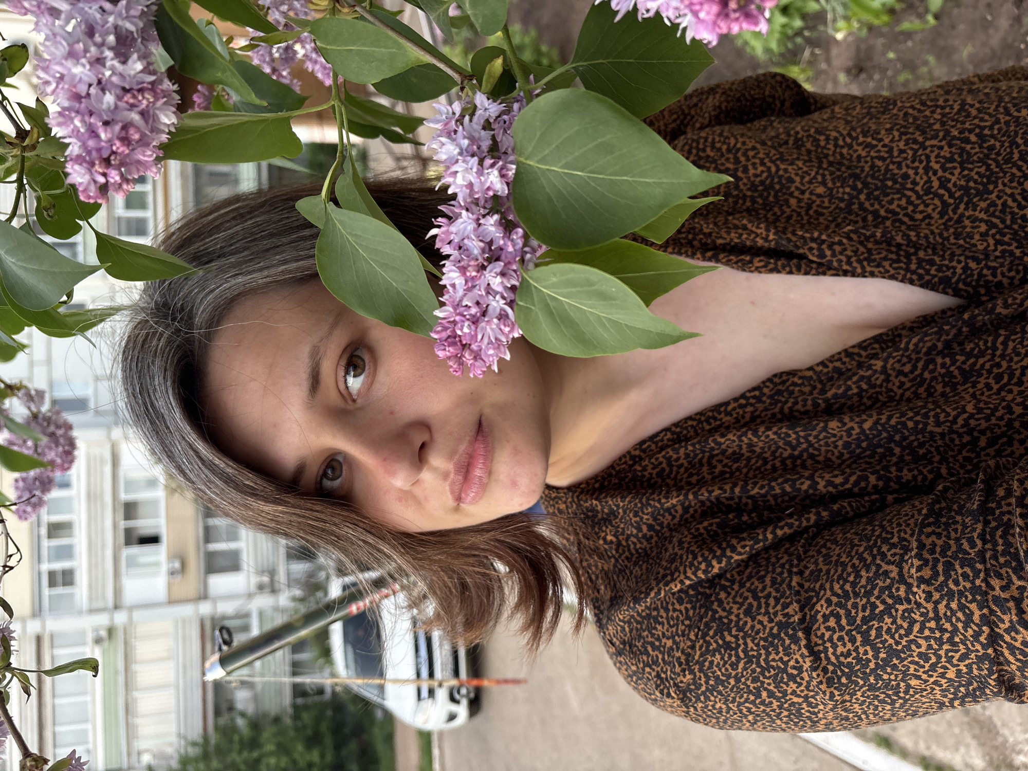 A woman with brown eyes and medium-length brown hair with streaks of grey is partially obscured by lilac blossoms. She is wearing a brown and black patterned top. A multi-story building is visible in the background. A white vehicle is parked on the street. The location is Chaikovsky, Russia.