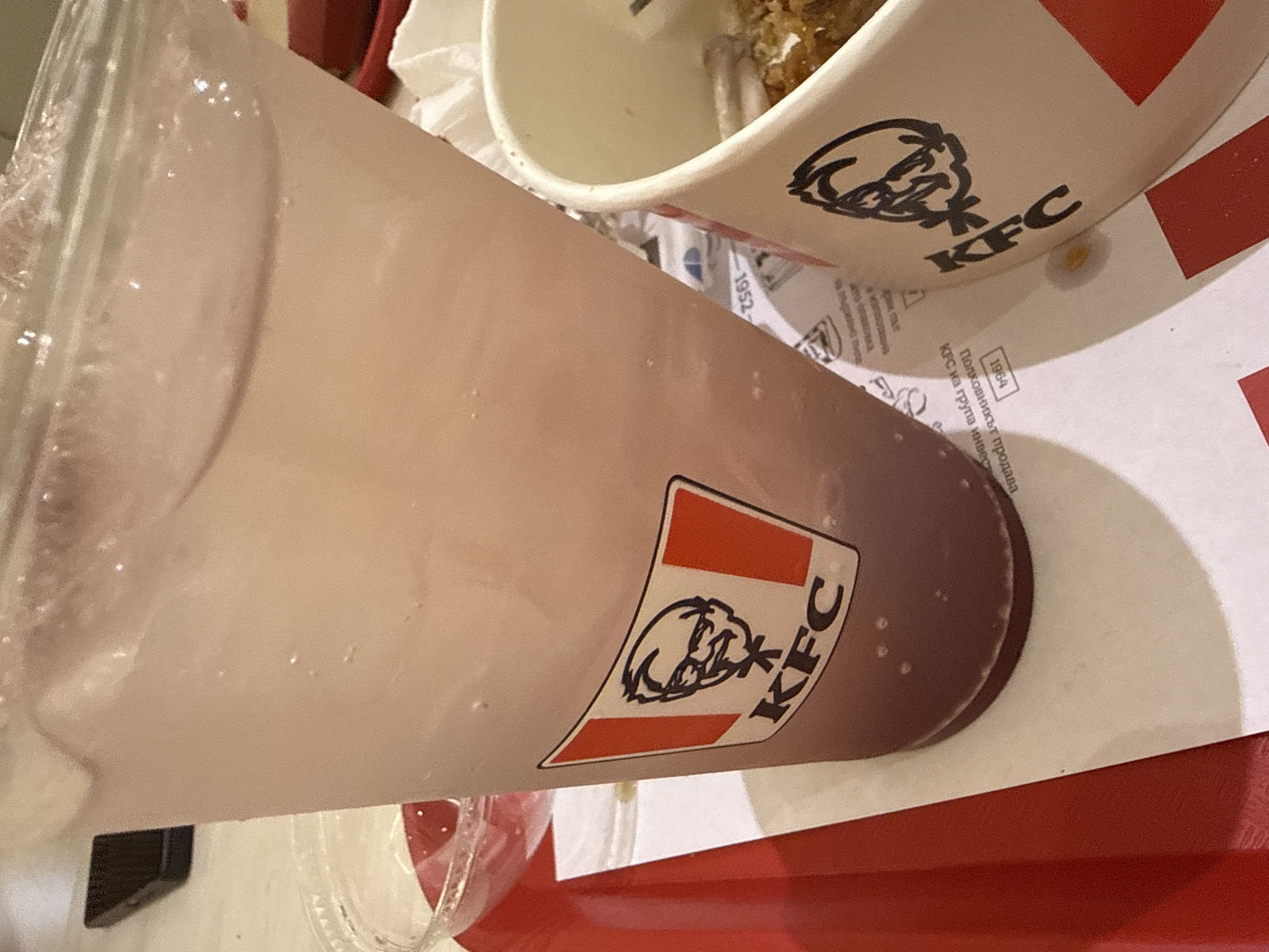 The image shows a KFC drink in a plastic cup and a KFC bucket on a red tray. There is a paper napkin and a sheet of paper with Cyrillic writing underneath the items. The location is likely a KFC restaurant in Plovdiv, Bulgaria, based on the language on the paper.
