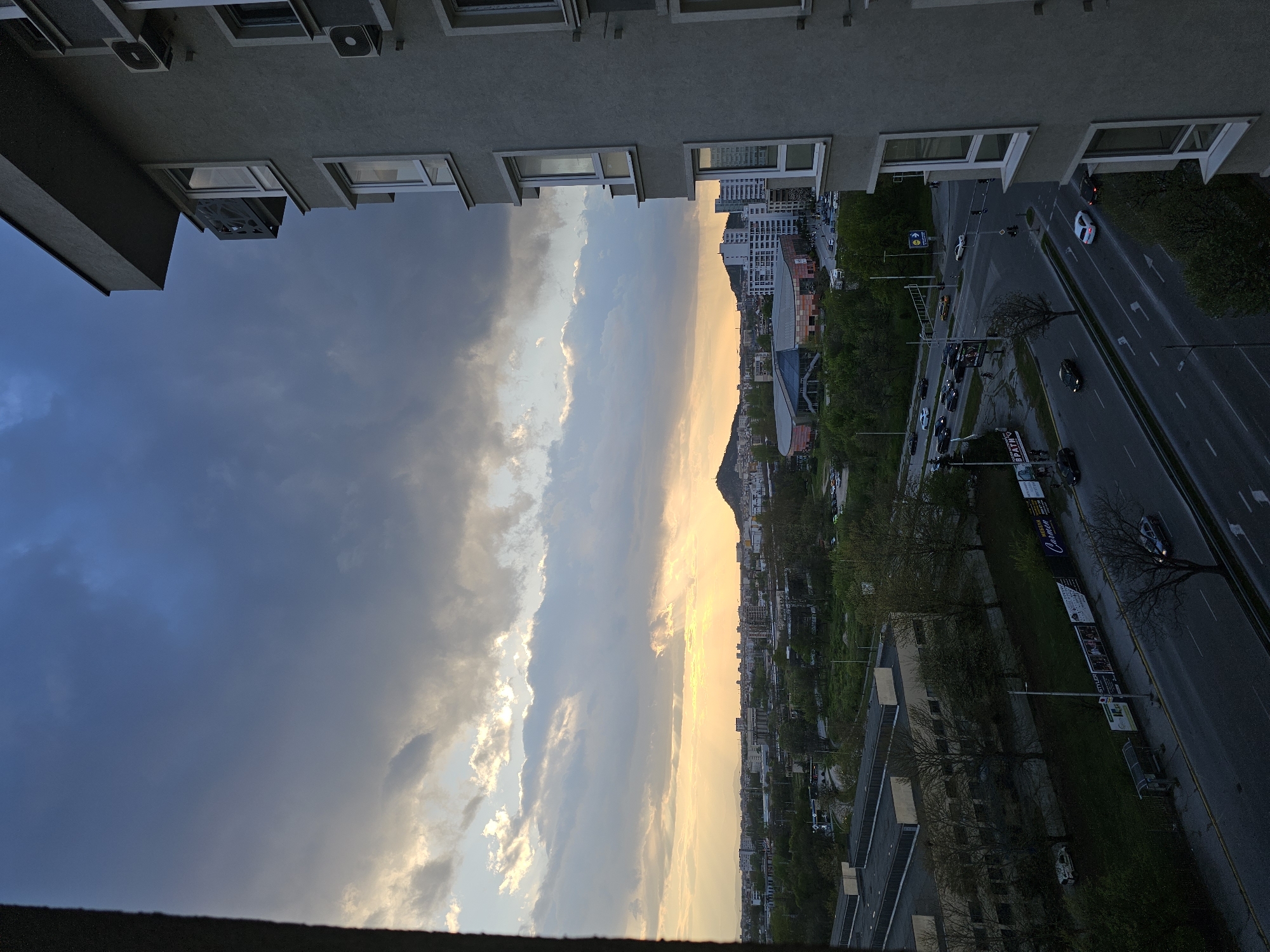 The image displays an urban landscape, likely Plovdiv, Bulgaria, captured from a high vantage point, possibly a window or balcony, during late afternoon or early evening. The scene is framed by parts of two buildings; the upper structure shows multiple windows and external air conditioning units, while a darker railing or edge of a building forms the lower border.
A prominent multi-lane road, lined with trees and streetlights, cuts through the middle ground. Cars are visible on the road, some appearing to be in motion, others parked. A traffic light and a blue road sign with a white arrow indicating "straight ahead" are discernible. The cityscape is dotted with various buildings of different heights and architectural styles, including one with a distinctive modern, curved facade. In the distance, a mountain or large hill rises against the horizon.
The sky is a dramatic focal point, exhibiting contrasting conditions. The left side is dominated by dark, heavy clouds, suggesting overcast weather, while the right side is bathed in the warm, golden and orange light of the setting sun, with sunbeams breaking through the clouds near the horizon. This creates a striking visual dichotomy of light and shadow across the sky.
Visible text includes "Carmen" on a large billboard next to the road, along with "STYLER" and "ОБУВКИ" (Bulgarian for "shoes"), advertising a commercial entity. Another sign partially displays "ПРАВНО" (Bulgarian for "legal"). The overall scene captures a vibrant urban environment under a dynamic and visually striking sunset.