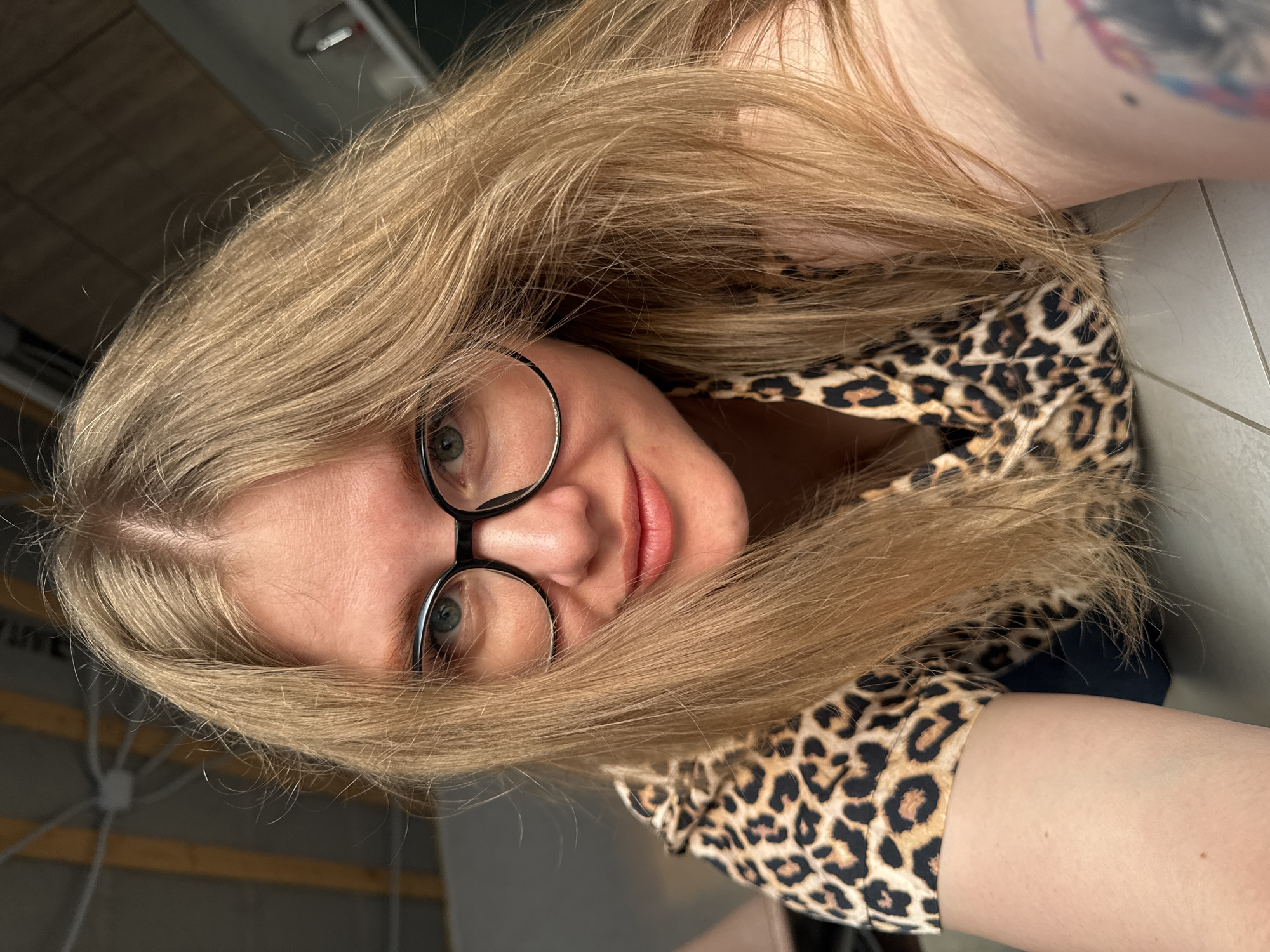 A woman with blonde hair and glasses is posing for a selfie. She is wearing a leopard print top. The location cannot be determined based on the image alone.