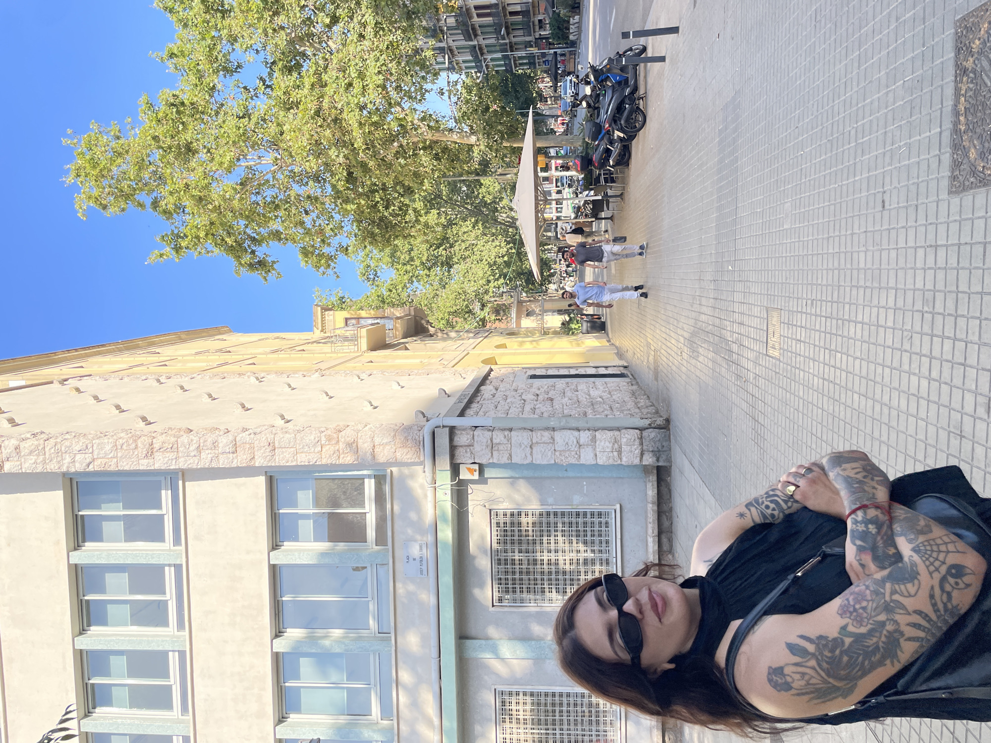 The image shows a woman with numerous tattoos on her arms, wearing sunglasses and a black top. She is seated on the sidewalk in what appears to be a European city. In the background, there is a building with windows, a tree with green foliage, and a street with parked motorcycles. The ground is paved with light-colored square tiles. There are other people walking in the background, some seated at tables under a large umbrella. The lighting suggests it is daytime, likely sunny, based on the bright sunlight and shadows. A sign on the building reads "PLAÇA DE JOSEP PLAJOAN DOMINGO". The overall scene suggests a casual urban environment in Barcelona, Spain.