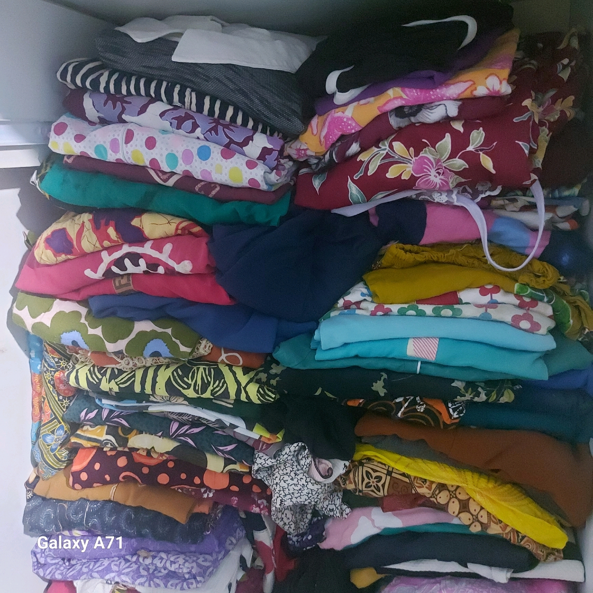 A top-down, medium shot captures a neatly folded stack of colorful clothes filling a drawer or shelf. The garments exhibit a variety of patterns and colors, including floral prints in shades of red, pink, and green, polka dots in blue, yellow, and pink, and abstract designs in black and white. Some of the fabrics are textured, while others appear smooth and silky. The clothes are arranged in distinct piles, creating a visually appealing and organized display. The overall impression is one of a well-kept wardrobe. The text "Galaxy A71" is overlaid on the bottom left of the image.