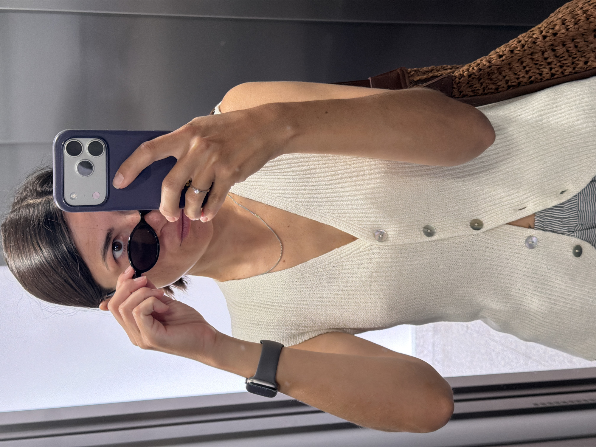 A woman is taking a selfie in a mirror. She is wearing a cream-colored, button-down cardigan, a silver necklace, and an Apple Watch. She is holding her sunglasses in one hand and her phone in the other. The phone case is blue. A straw bag is visible in the upper right corner of the image. The location appears to be an indoor space with reflective surfaces. There is a window or opening behind her with horizontal bars. The location is not confirmed to be in Budva, Montenegro.