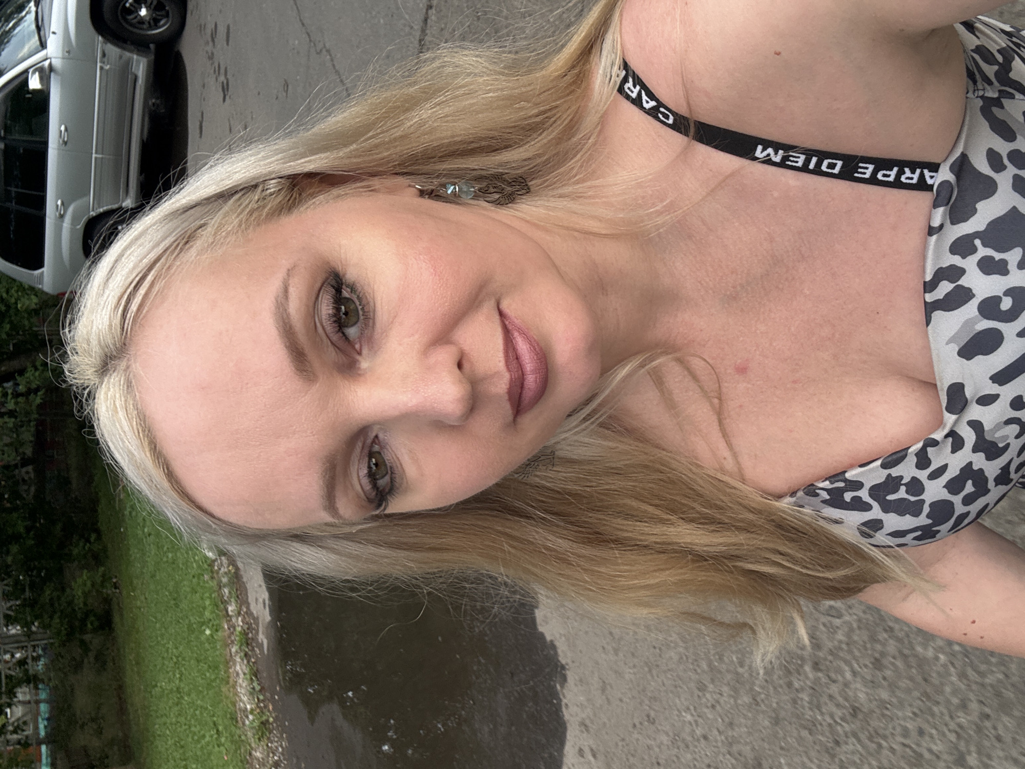 A blonde woman in a leopard print top stands in an urban outdoor setting. She is looking at the camera, and her face is the main focus. She has makeup on and is wearing an earring on her right ear. Behind her, a silver SUV is parked on the street, and there is a grassy area with trees in the background. A puddle of water is visible on the asphalt in the foreground. The image appears to be taken during the daytime, possibly in Komsomolsk-on-Amur, Russia, as indicated by the context.