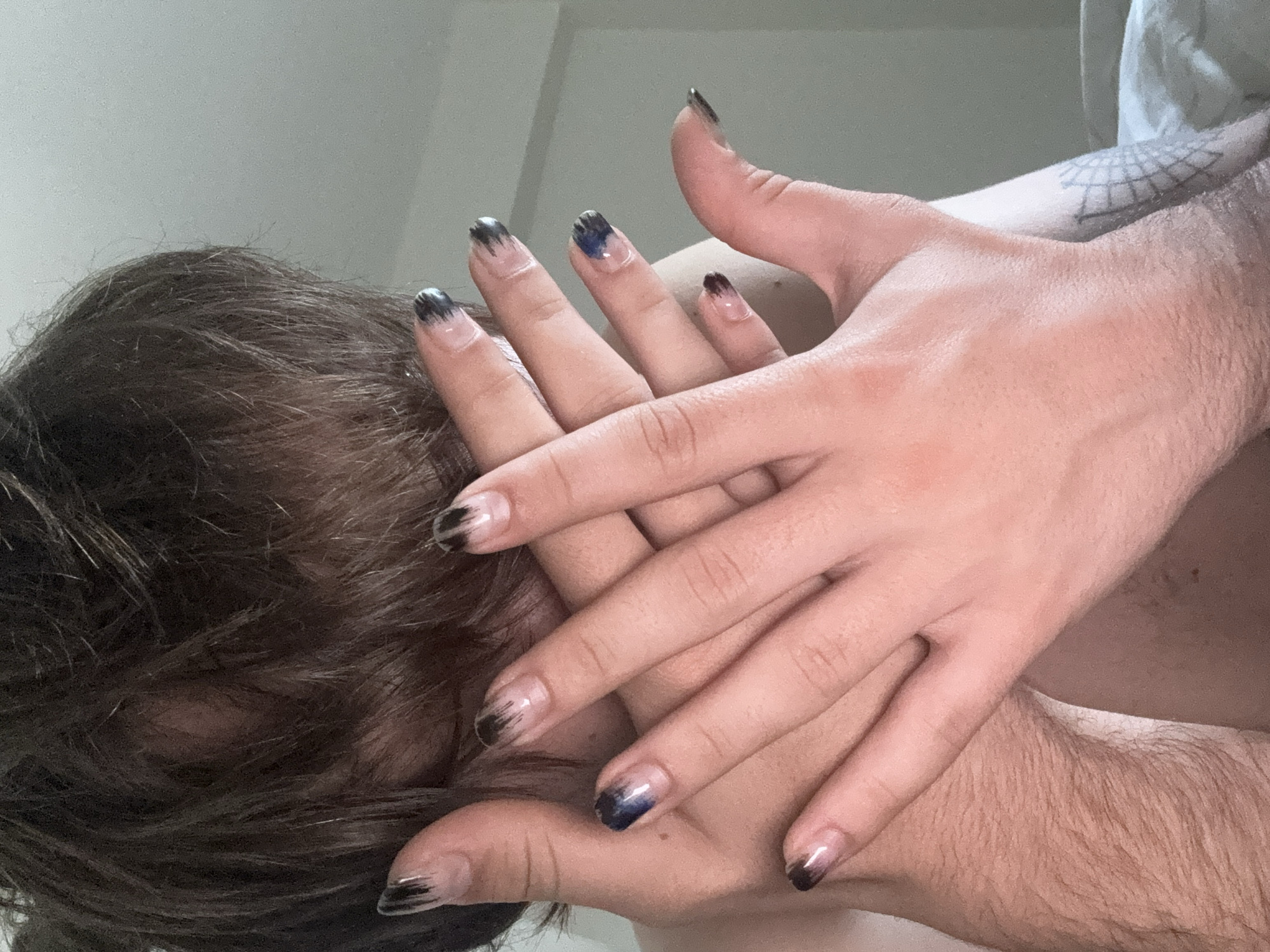A close-up, low-angle shot shows a person with brown hair partially visible in the lower-left of the frame. Two hands with fingernails painted in an ombre style, transitioning from clear at the base to black at the tips, are intertwined. The hands are pale, with visible veins and fine hairs. The right hand rests on the shoulder of a person with a tattoo visible on the right shoulder. The background is a light, neutral color, possibly a wall. There is no specific indication of location in Tel-Aviv, Israel.