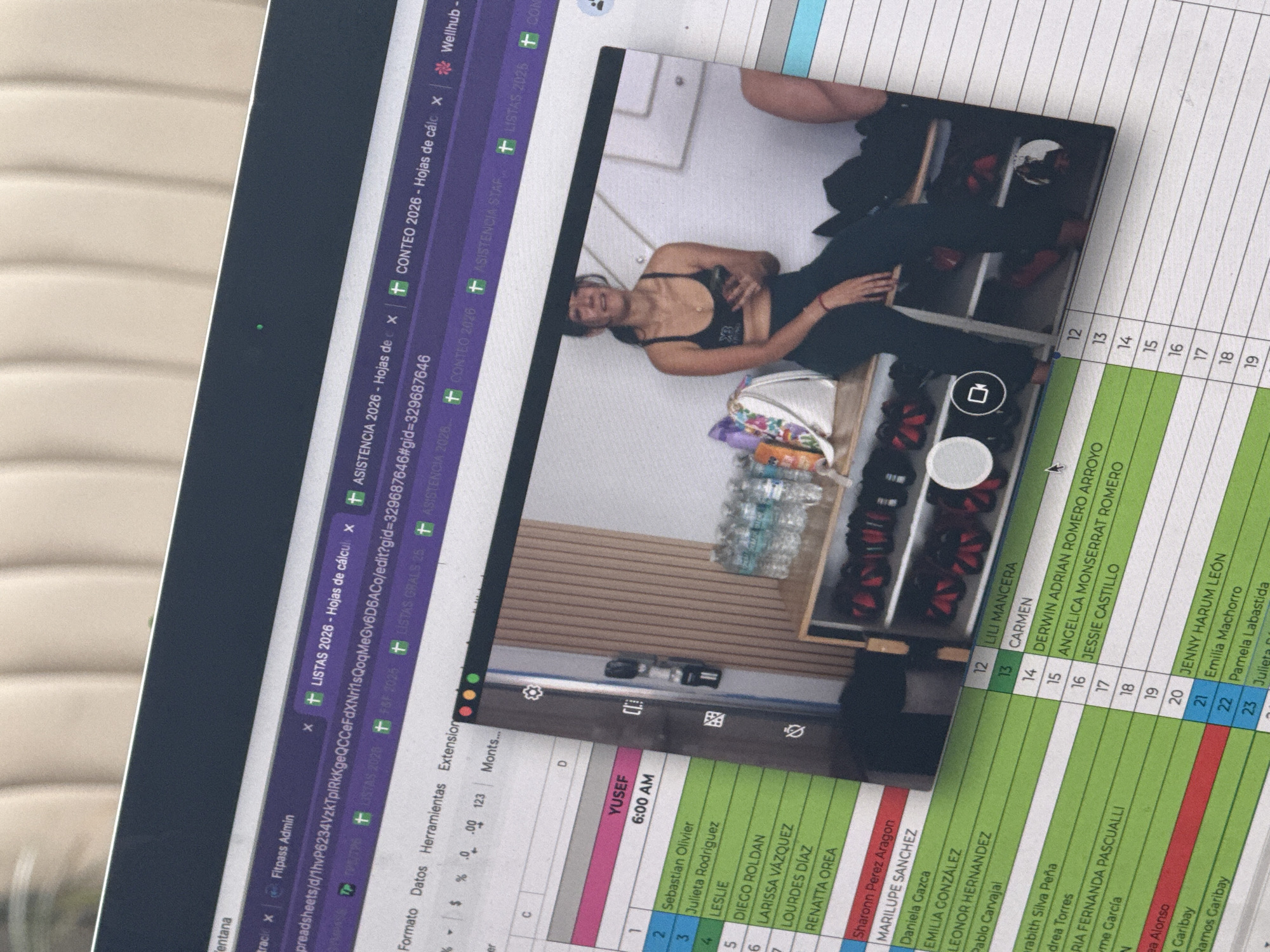 The image shows a photograph of a computer screen displaying a spreadsheet and a photo of a person. The spreadsheet appears to be an attendance sheet or similar record with names and times (6:00 AM). The photo within the screen shows a woman in athletic attire (sports bra and leggings) holding her phone in a locker room, with rows of lockers, water bottles, and what appear to be boxing gloves or similar equipment in the background. Based on the spreadsheet titles "LISTAS 2026," "ASISTENCIA 2026," and "CONTEO 2026," the scene could be related to a fitness class or gym check-in. Puebla, Mexico is not mentioned in this image.