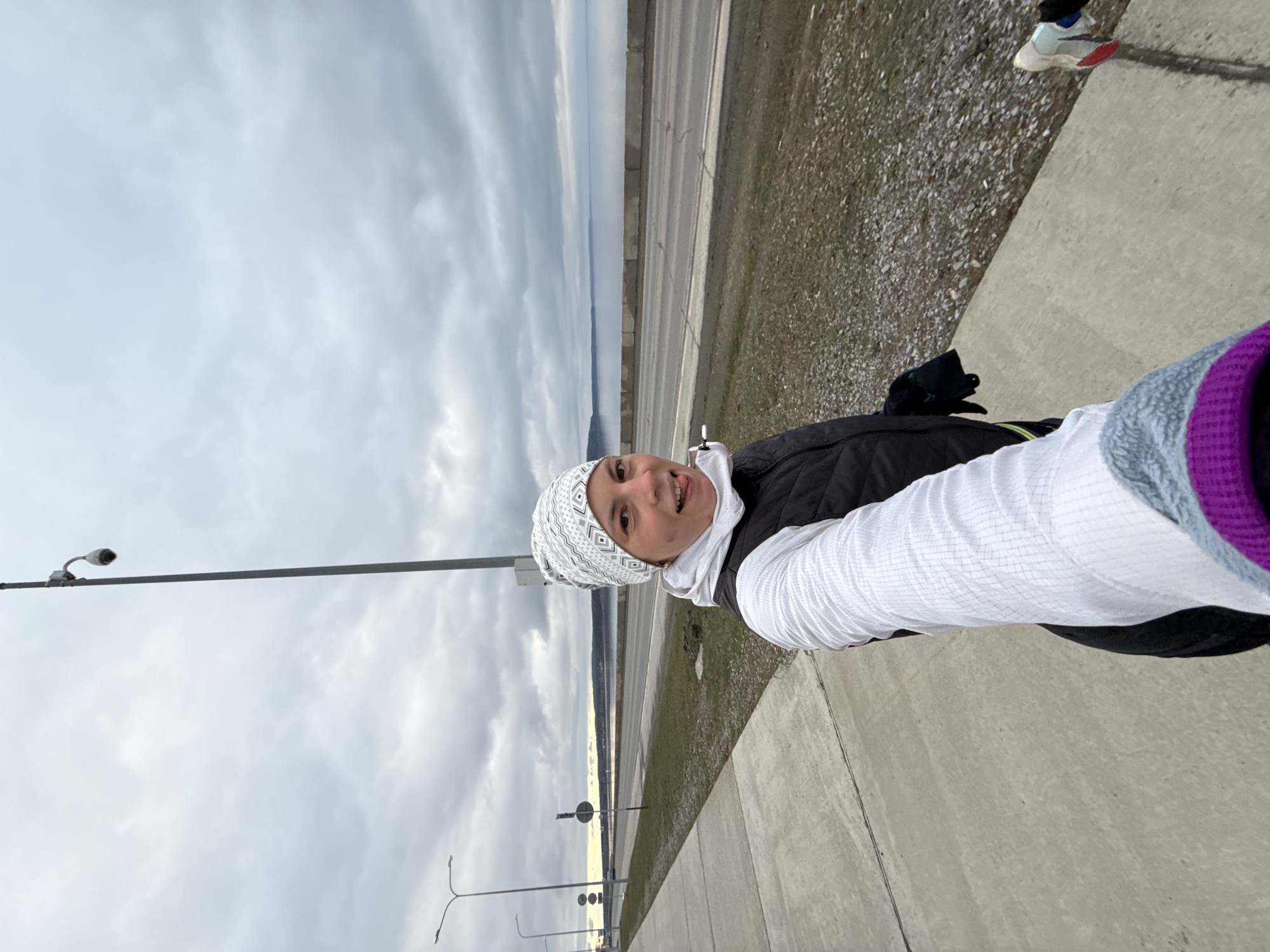 A selfie portrait captures a woman smiling playfully, with her tongue slightly extended, against an outdoor backdrop on an overcast day in Chaikovsky, Russia. The woman, positioned on the right side of the frame, is wearing a white patterned beanie, a white head covering (likely a hijab or neck gaiter), a black puffer vest, and a white long-sleeved shirt with a textured, possibly grid-like pattern.
She stands on a concrete sidewalk, which extends across the foreground. To the left of the sidewalk is a gravel and dirt shoulder, leading to a multi-lane highway or large road. A concrete barrier separates the road from the wider landscape. Beyond the road, a vast body of water, likely the Kama River or Votkinsk Reservoir, stretches towards a distant, low-lying landmass or hills on the horizon. The sky is largely grey and cloudy, indicating overcast weather.
Visible objects in the scene include a tall metal light pole with a security camera mounted at its top, positioned on the far left. Further down the road, another light pole with multiple arms and a small, circular sign are also present. The lighting is diffused, suggesting a lack of direct sunlight, consistent with the cloudy conditions. The woman's warm attire implies cool or cold temperatures. No distinct text is visible anywhere in the image.