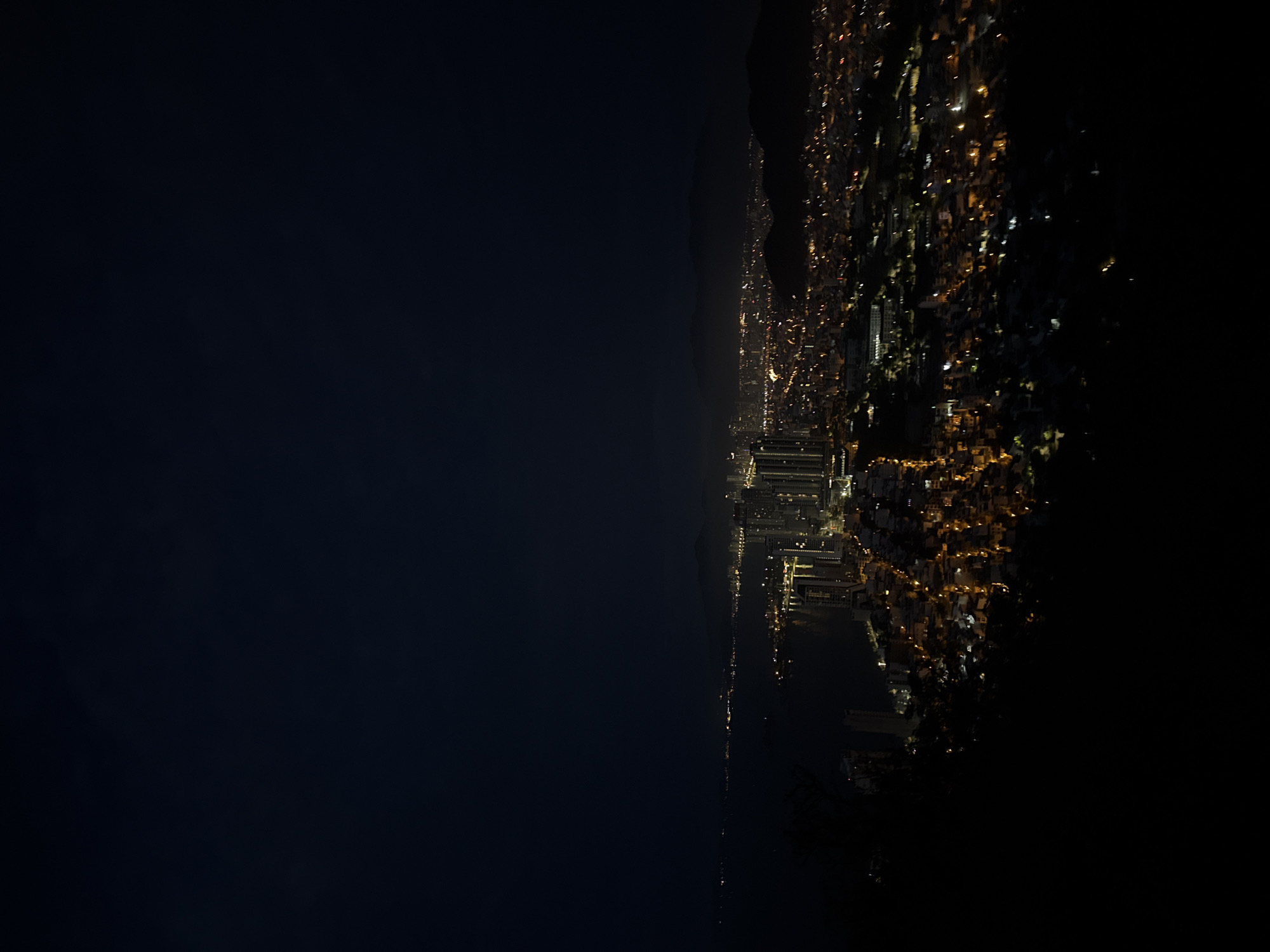 A dark, low-angle nighttime view of a city, likely Nha Trang, Vietnam, featuring building lights and mountainous terrain in the distance. The left portion of the image is a large expanse of dark, likely representing water or sky. The right portion features densely packed buildings with glowing lights, stretching from the foreground into the distance. A line of mountains silhouettes against the night sky further inland. The foreground appears to be a dark, unlit area, possibly vegetation.