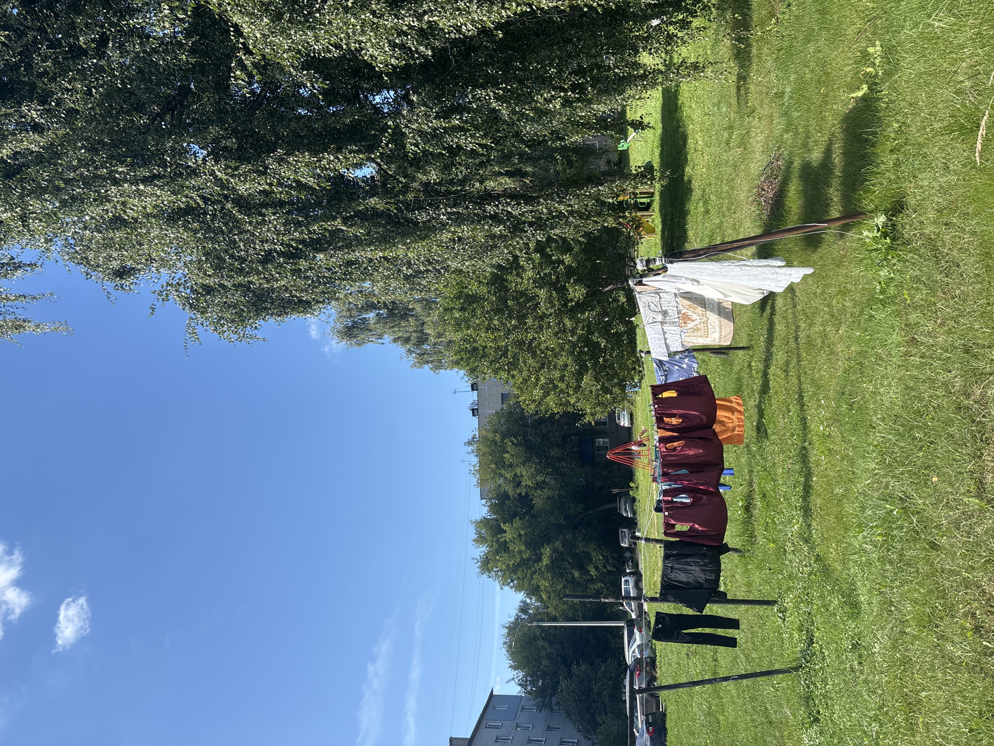 The image depicts an outdoor scene in Kstovo, Russia, on a clear, sunny day with a bright blue sky and scattered white clouds. In the foreground, laundry is drying on a clothesline strung between two poles. The laundry includes various items of clothing such as black pants, a black jacket, maroon shirts or vests, and orange garments, as well as white fabric and patterned linens. The clothesline is supported by rustic, metal poles set in a grassy area.
Behind the clothesline, a lush green lawn stretches towards a row of trees and buildings. A large, mature birch tree with its characteristic white bark and dense foliage dominates the right side of the frame, casting shadows on the ground. In the mid-ground, several apartment buildings, typical of Soviet-era architecture, are visible, interspersed with more trees. Cars are parked along the side of one of the buildings. A children's playground with a red frame and yellow and green structures can be seen in the distance, nestled amongst the trees. The overall impression is a residential area in Kstovo, Russia, with everyday life activities taking place outdoors.