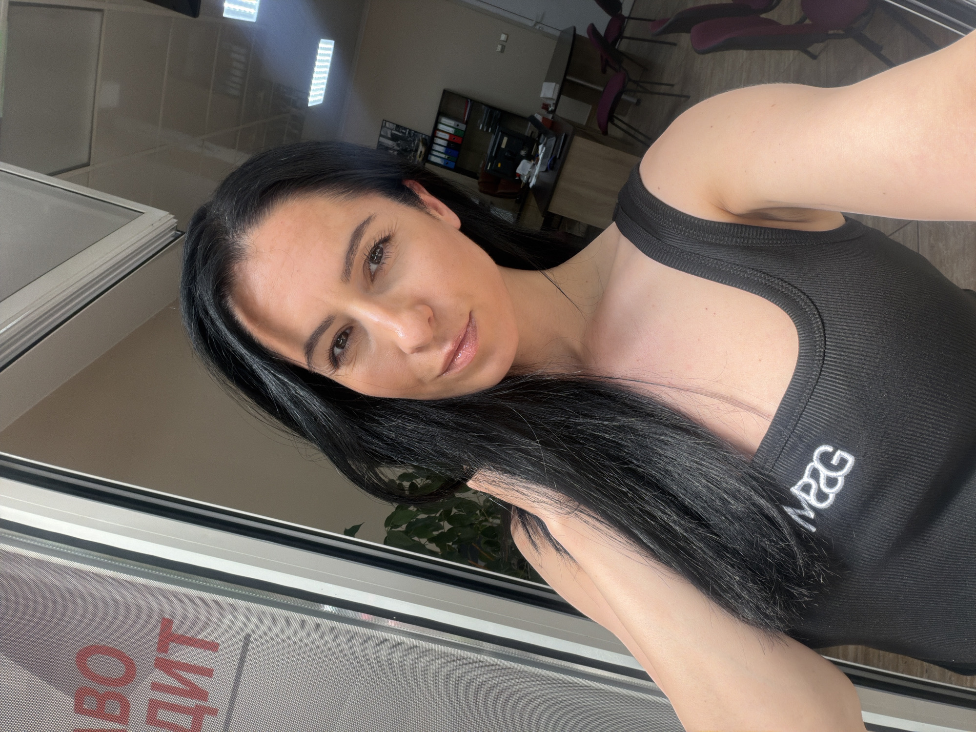 A selfie of a woman with dark hair is taken in an indoor setting. She is wearing a black tank top with white lettering on the chest. The background shows office furniture, including a desk with chairs and filing cabinets. A window is visible on the left with writing in a non-English alphabet displayed on the window screen. The location may be in Pleven, Bulgaria.