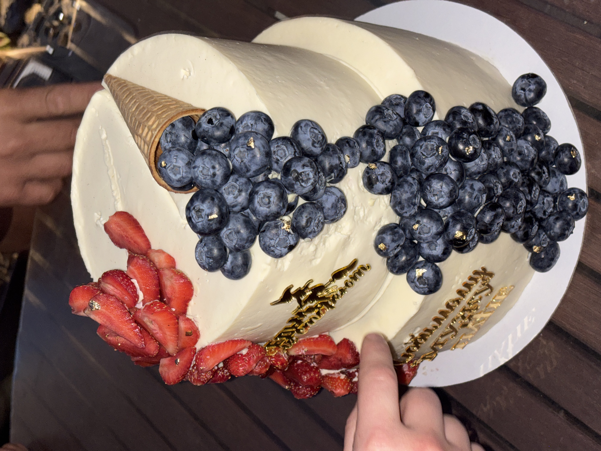 A two-tiered white frosted cake is decorated with fresh strawberries, blueberries, and edible gold leaf. A waffle cone is embedded in the top tier, spilling blueberries onto the side. Golden text is affixed to both tiers of the cake. A person's hands are visible in the foreground, one touching the cake. The cake is set on a white platter and sits on a wooden surface. The setting appears to be outdoors, with a dark background suggesting evening. This image is not tagged with a specific location in Barnaul, Russia.