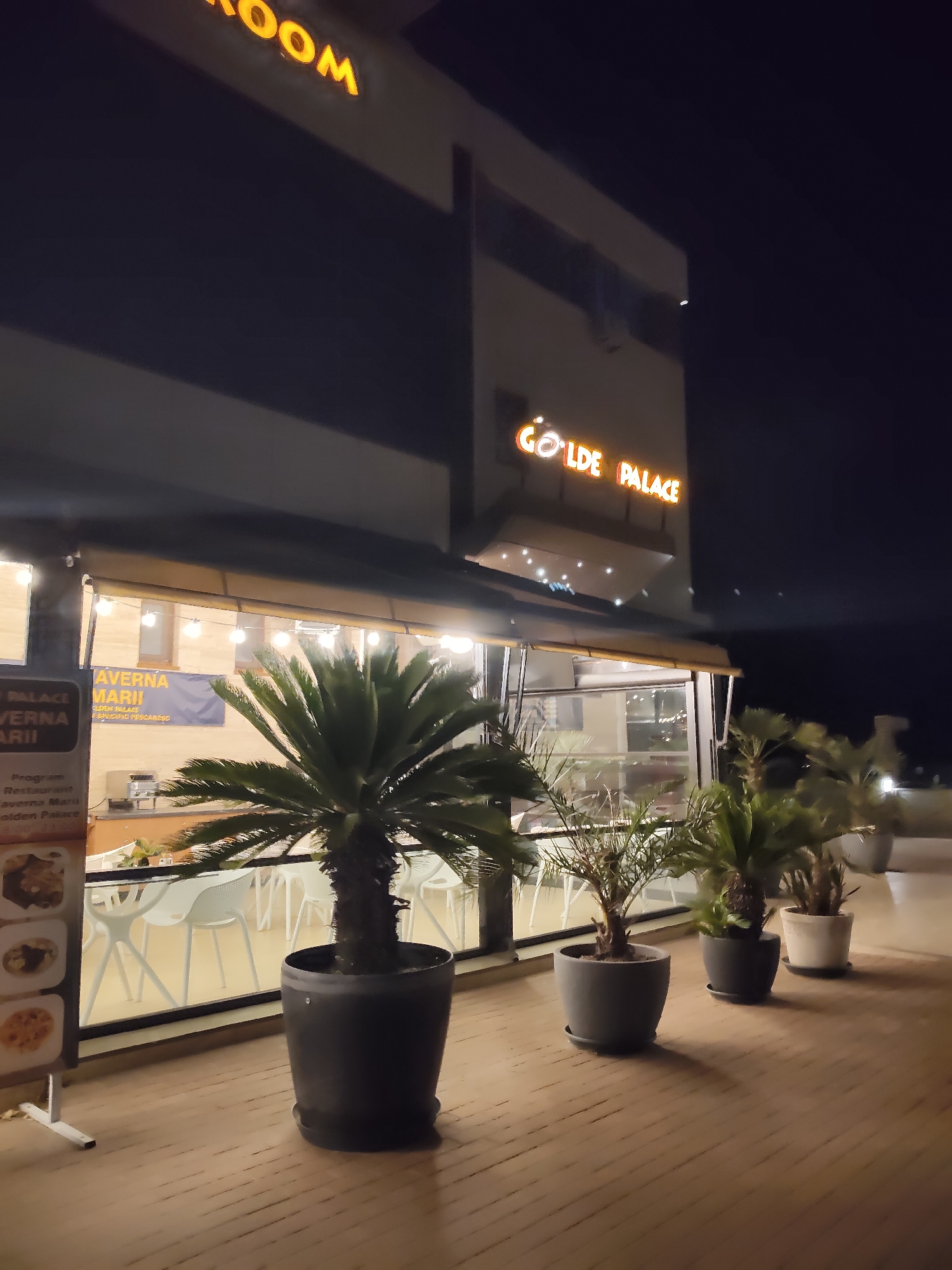The image shows a nighttime view of the "Golde Palace" restaurant and banquet hall located in Constanța, Romania. The establishment features illuminated signage in orange neon letters. In the foreground, several potted palm plants are arranged on a brick-patterned outdoor flooring. Behind the plants, a glass-enclosed seating area of the restaurant is visible, with white chairs and tables set up inside. String lights are hung above the entrance to the establishment. A vertical sign board with menu item images is positioned to the left of the frame. The overall scene suggests an outdoor dining or event space at night.