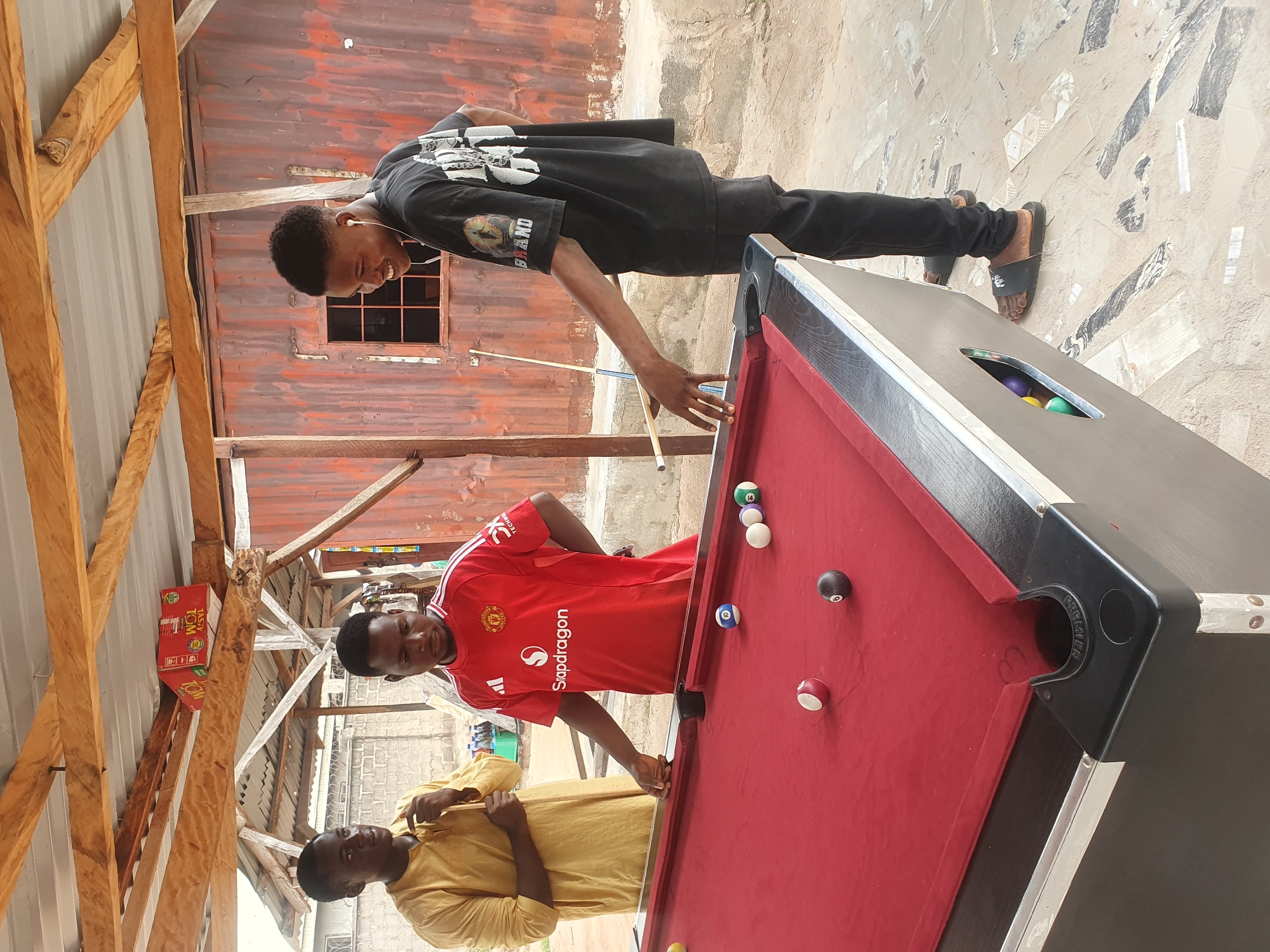 Three young men are playing pool in an outdoor, covered area in Lokoja, Nigeria. The structure appears to be a makeshift shelter with a corrugated metal roof supported by wooden beams and poles. The walls are a combination of corrugated metal and concrete block. In the background, a weathered red corrugated metal structure with a small, barred window is visible. The ground is a mixture of sand and patterned tiles.
Two of the men are actively engaged in the pool game. One man, wearing a yellow tunic, stands on the left, holding a pool cue. Another man in a red Manchester United jersey stands by the table, his hand near a pool cue, looking down at the balls. The third man, wearing a black t-shirt and black pants, stands to the right, holding a pool cue and smiling. He is wearing earphones.
The pool table is covered with red felt and has several balls on it, including a black 8-ball, a red and white striped ball, a numbered blue ball, and other colored balls. The balls are positioned on the table and some appear to be in pockets. Pool cues are leaning against the table and being held by the players. There is a red and white box with lettering, possibly a food item, placed on a wooden beam above the players. In a compartment on the side of the pool table, several colored pool balls are visible.