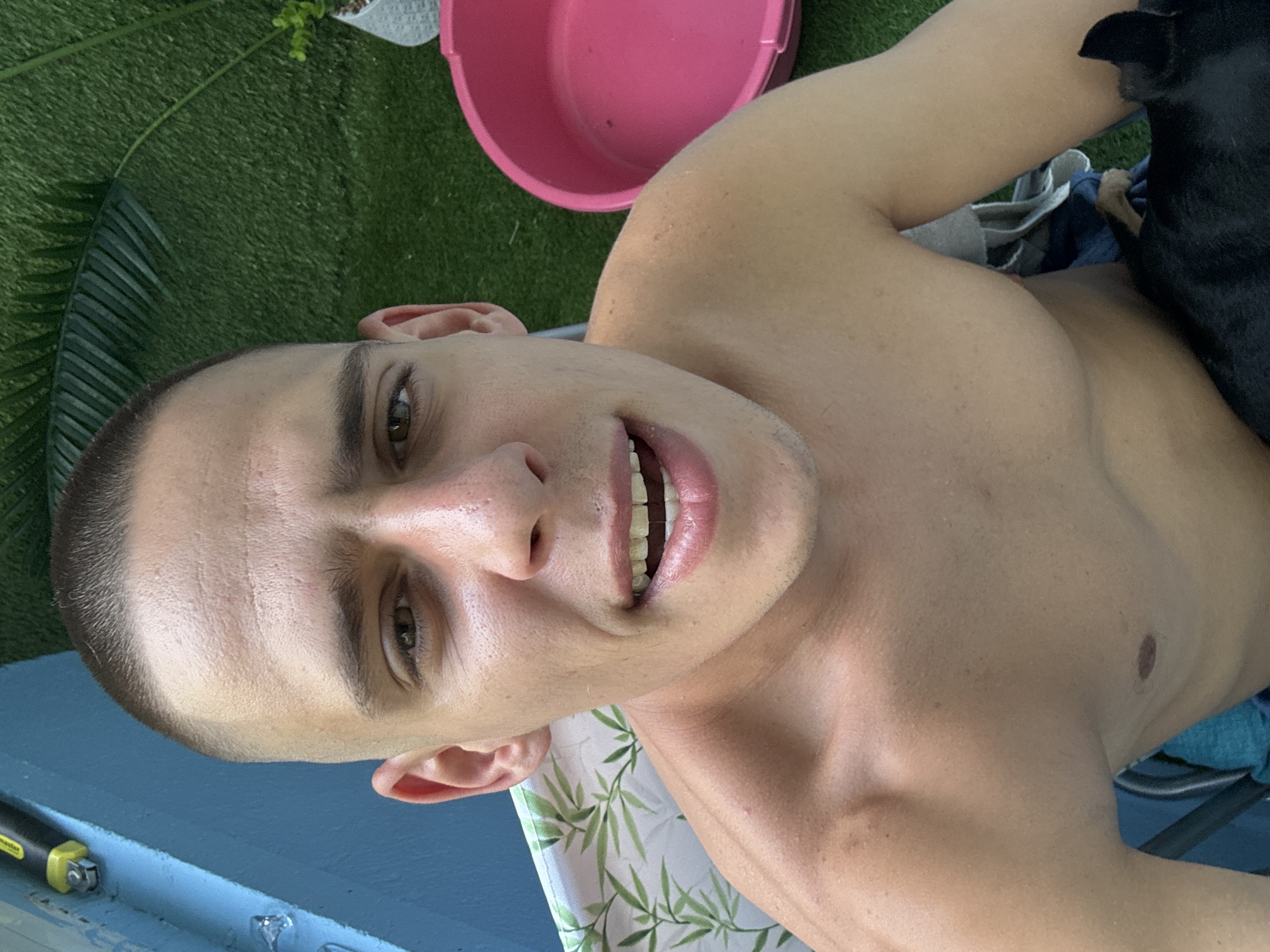 The image shows a close-up of a man from the chest up, lying on his back. He has a buzz cut and appears to be outdoors, possibly in a garden or yard, given the artificial grass and a large pink basin in the background. A black dog is partially visible in the upper right corner, nuzzling against the man's shoulder. The man's mouth is open, as if he is smiling or speaking, and his eyes are looking directly at the camera. He is wearing a light-colored towel with a green leaf pattern. A blue surface, possibly a table or lounge chair, is visible in the lower part of the frame, with a yellow and black tool resting on it. The lighting suggests it could be daytime, possibly a warm day, given the man is shirtless. There is no visible text in the image.