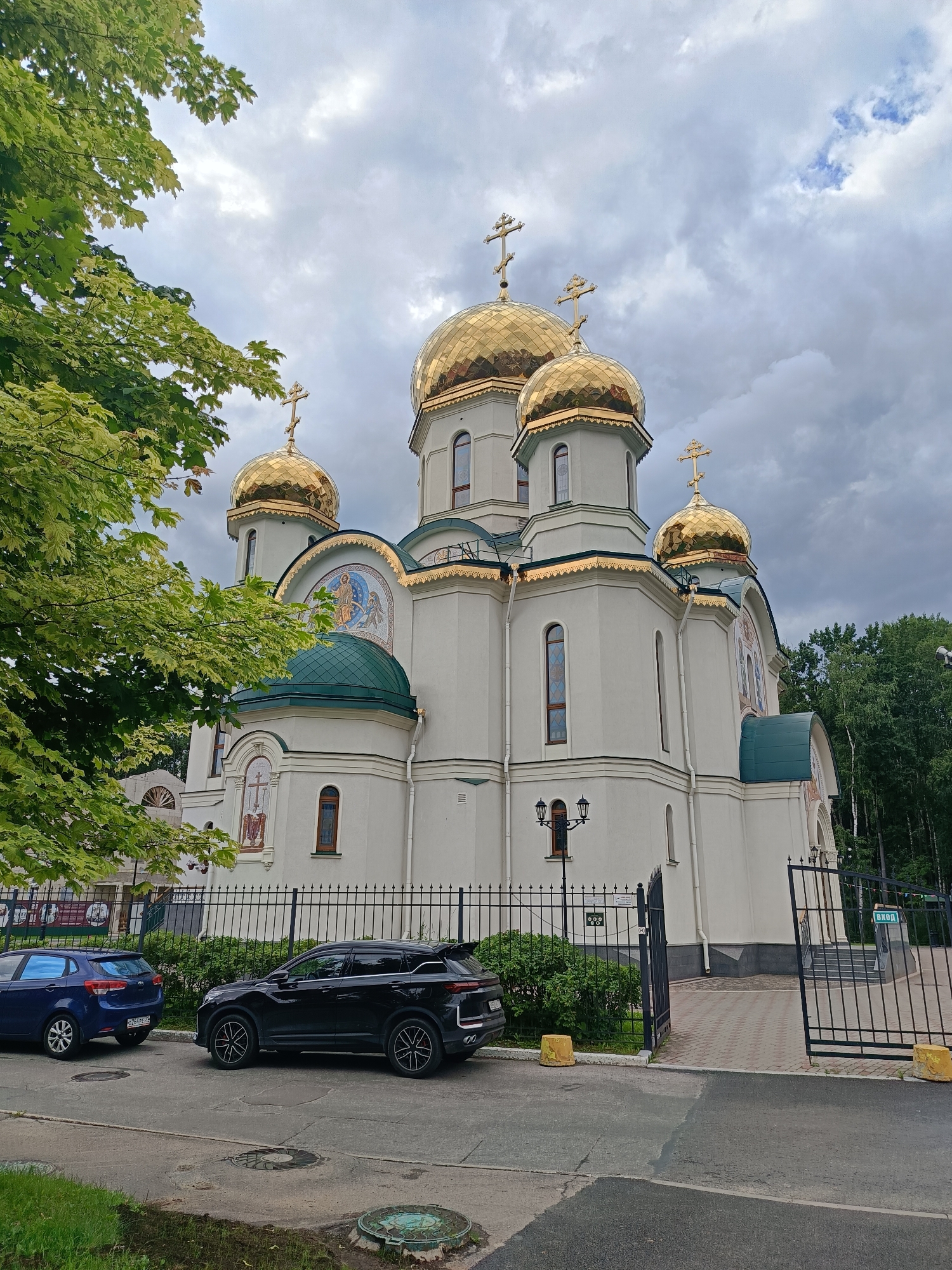 This is a photograph of the Church of the Resurrection of Christ, located in the city of Saint Petersburg, Russia. The church is a prominent building with several golden domes topped with crosses. The facade is white with green accents on some of the roofs. There are mosaic images on the exterior walls. In front of the church, there is a black metal fence with an open gate. Two cars are parked on the street in front of the fence: a blue hatchback and a black SUV. Trees and greenery surround the church and the street. The sky is cloudy, indicating an overcast day.