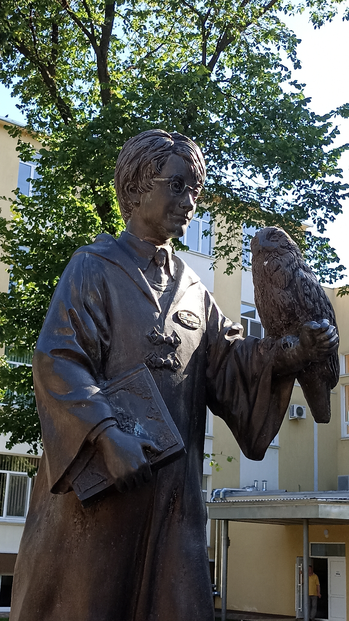A bronze statue of a young person wearing round glasses and a robe stands in front of a yellow building. The statue holds a book in its left hand and an owl perched on its right arm. The statue is located in Tiraspol, Moldova, in front of the Taras Shevchenko Transnistrian State University. A person wearing a yellow shirt and grey pants is visible in the doorway of the building in the background. The scene is illuminated by sunlight, casting shadows on the statue and surrounding area.