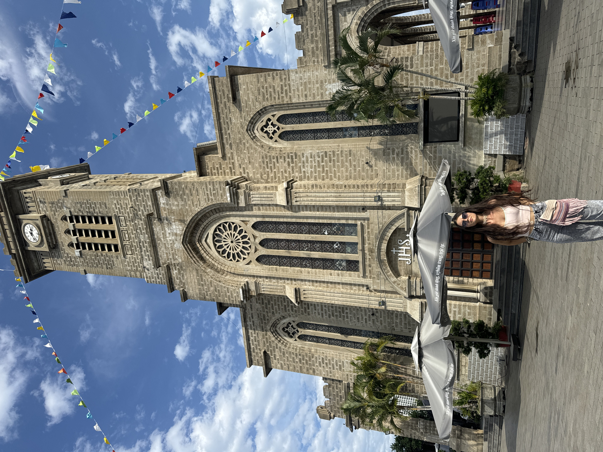 A young woman with long, dark hair and sunglasses stands centered in the foreground, facing the camera. She is wearing a light pink tank top and distressed denim jeans, with a fringed crossbody bag slung over her shoulder. Behind her, the imposing stone facade of the Nha Trang Cathedral, also known as the Stone Church, dominates the frame. The cathedral features Gothic architectural elements, including arched windows, a rose window above the main entrance, and a tall bell tower with a clock. Colorful triangular flags are strung across the sky above the church. Several large gray umbrellas are set up in front of the cathedral, some displaying text in Vietnamese. Palm trees and potted plants are visible around the entrance area. The ground in front of the cathedral is paved with stone tiles. The sky is blue with scattered white clouds. The scene appears to be a public square in front of a prominent religious landmark.