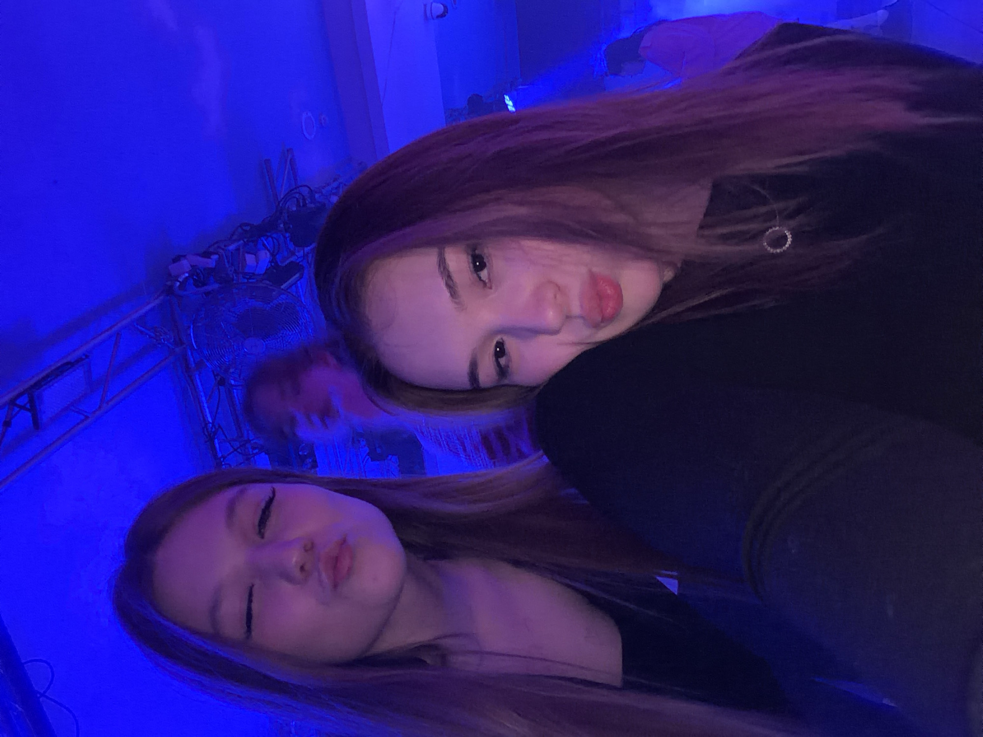 Two young women are posing closely for a selfie. The woman on the left is slightly lower in the frame, pouting her lips toward the camera. The woman on the right is leaning in from above, also posing with pursed lips. Both have long hair. The lighting is predominantly blue, suggesting an indoor setting, possibly a club or event space. Behind them, there's a glimpse of a stage-like structure and some equipment. In the background, a partially visible figure is blurred. The exact location in Chaikovsky, Russia, cannot be definitively determined from the visual details provided in the image.