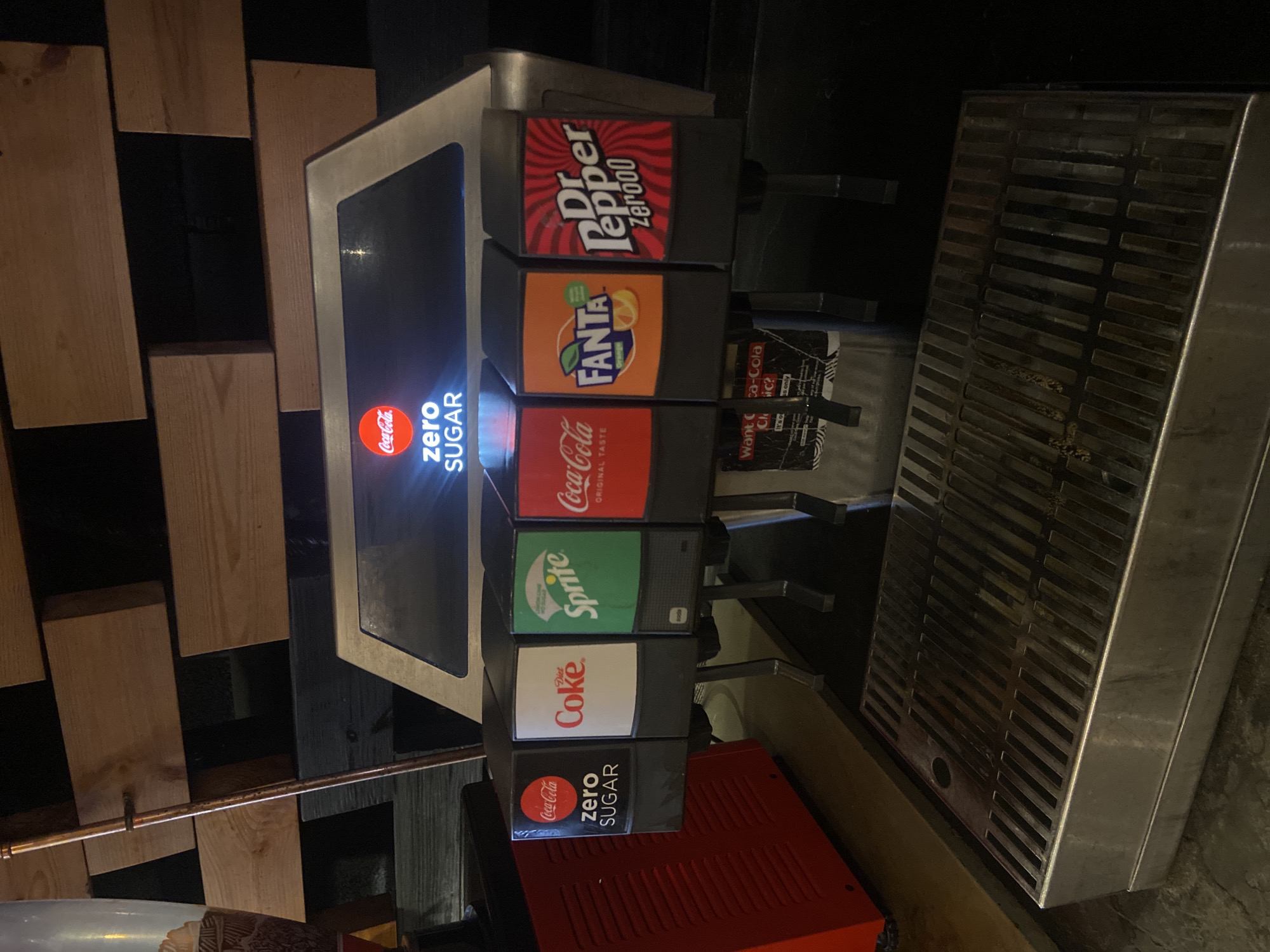 The image shows a soda fountain machine with various drink options. The top row displays Dr. Pepper Zero00, followed by Fanta. The middle row features Coca-Cola (original taste), Sprite, and Coca-Cola Zero Sugar. The bottom row has the Coca-Cola Zero Sugar option. Next to the soda fountain is a stainless steel drain tray. The soda machine is located indoors in what appears to be a food service establishment. The backdrop includes a textured wall with various tones of wood. There is a pipe running below the wood wall. The location appears to be in a food service establishment and there is no evidence to indicate that it is located in London, United Kingdom.