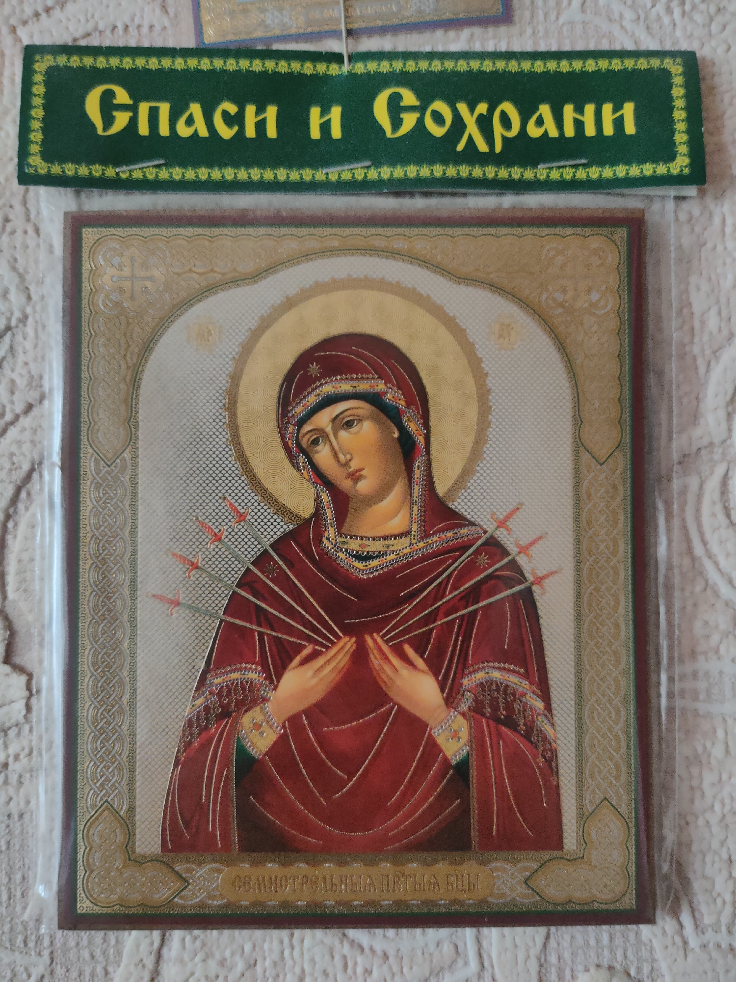 An Eastern Orthodox icon of the Virgin Mary, pierced by seven swords, is displayed against a white, textured background. The icon is framed with ornate gold detailing and has a dark brown border. Above the icon, a green ribbon with the inscription "Спаси и Сохрани" (Save and Protect) in yellow Cyrillic lettering is attached. The item appears to be a religious souvenir, possibly purchased or displayed in Oktyabrsky, Russia, given the cultural context of Eastern Orthodox iconography and the inscription. There are no people visible in the image.