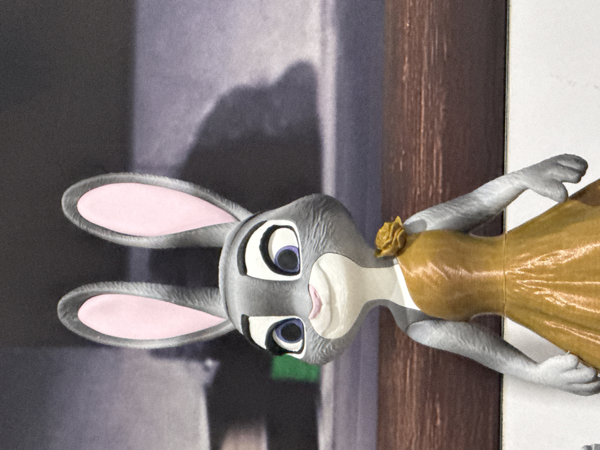 This is a close-up, eye-level, medium shot of a Judy Hopps action figure from Disney's Zootopia. The figure is depicted from the upper chest up, with its head slightly tilted to the left. Judy Hopps is wearing a gold, one-shouldered dress with a rose accent on the shoulder. The figure has large, expressive blue eyes, pink ears, and a gray furry body. The background is out of focus and appears to be a mural or a large photograph depicting a city skyline at dusk, possibly in Bangkok, Thailand, given the presence of characteristic skyscrapers and the warm, diffused lighting. The lighting on the figure is soft and diffused, highlighting the texture of the plastic. There are no people or animals present other than the action figure.