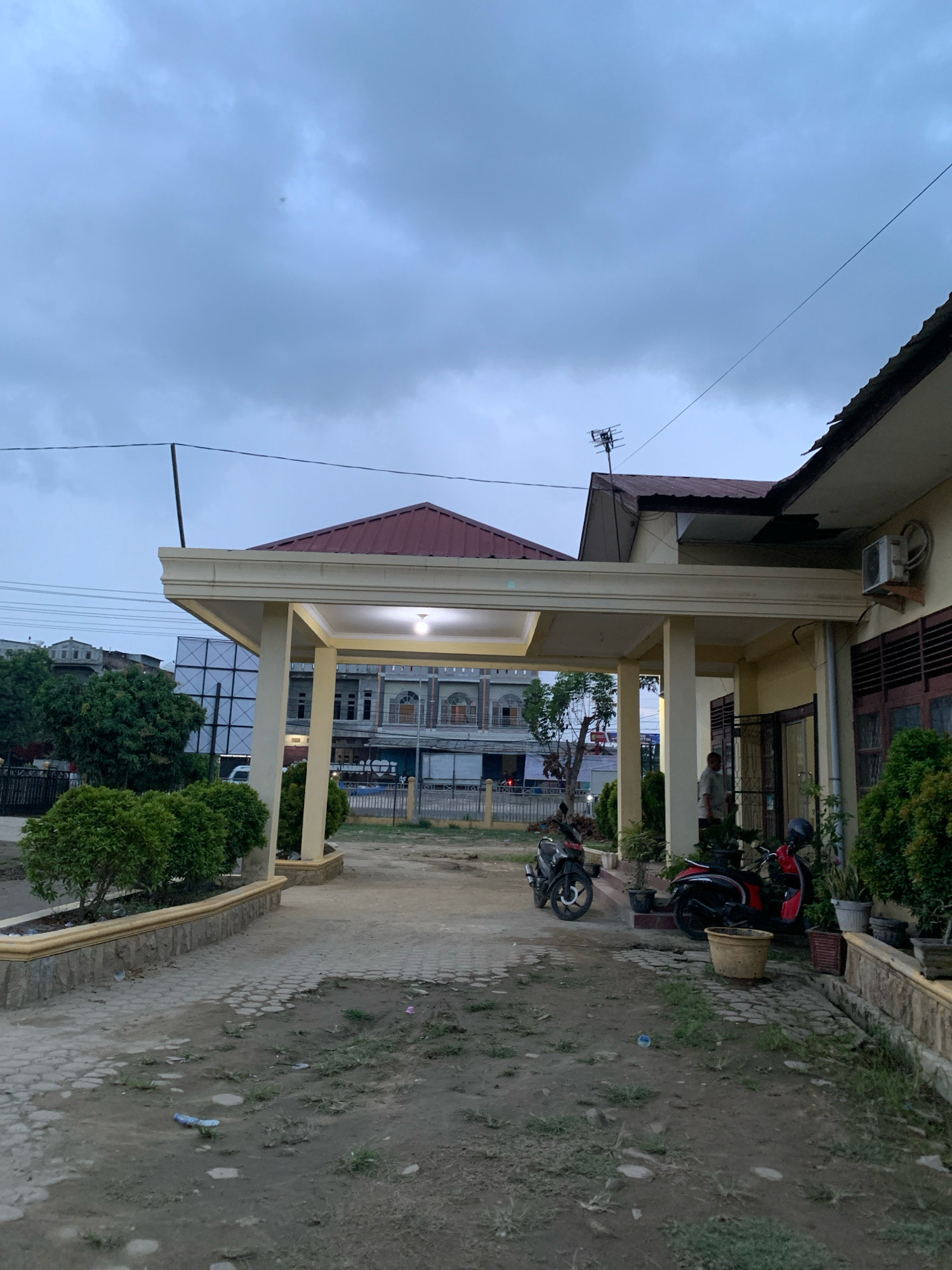 The image depicts a covered walkway in front of a building, likely a residence or small office, in Pantegurah, Indonesia. The sky is overcast with a dull, gray hue, suggesting it might be early morning or late afternoon. A prominent feature is the yellowish-beige covered walkway supported by pillars, leading to the entrance of the building. The building itself is painted a light yellow and has dark brown window frames. To the left of the walkway, there are manicured green bushes planted in raised beds with stone borders.
In the foreground, the ground is a mix of dirt, gravel, and some patchy grass. Several pieces of litter are scattered on the ground, including what appears to be a plastic bottle and a candy wrapper. Two motorcycles are parked under the covered walkway: a black scooter on the left and a red and black scooter on the right. A man in a light-colored shirt and dark pants is standing near the entrance of the building, conversing with someone who is not fully visible.
In the background, across from the building, there are other structures. One building appears to be a multi-story commercial or institutional building with arched windows and a red roof. Further in the distance, there are more buildings and trees, with some visible power lines and a large white billboard with dark text. An antenna is mounted on the roof of the building to the right, and an air conditioning unit is visible on its exterior wall. The overall impression is a mundane, everyday scene in a semi-urban or suburban setting.