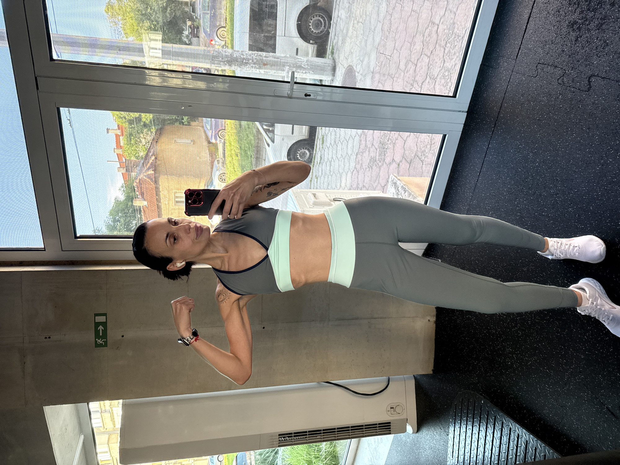 A woman is posing for a selfie in a gym. She is wearing athletic clothing, including a gray sports bra with mint green accents and matching leggings, and white sneakers. She is holding a smartphone in her left hand, and her right arm is flexed in a bicep pose. She has dark hair pulled back and is wearing earbuds.
The background shows a concrete wall with a green emergency exit sign, a white wall-mounted heater, a large glass window, and a black rubber floor. Outside the window, a building and a parked van are visible.
Based on the details visible in the image, including the building's architecture and the make of the parked van, the location is likely the city of Pleven, Bulgaria.