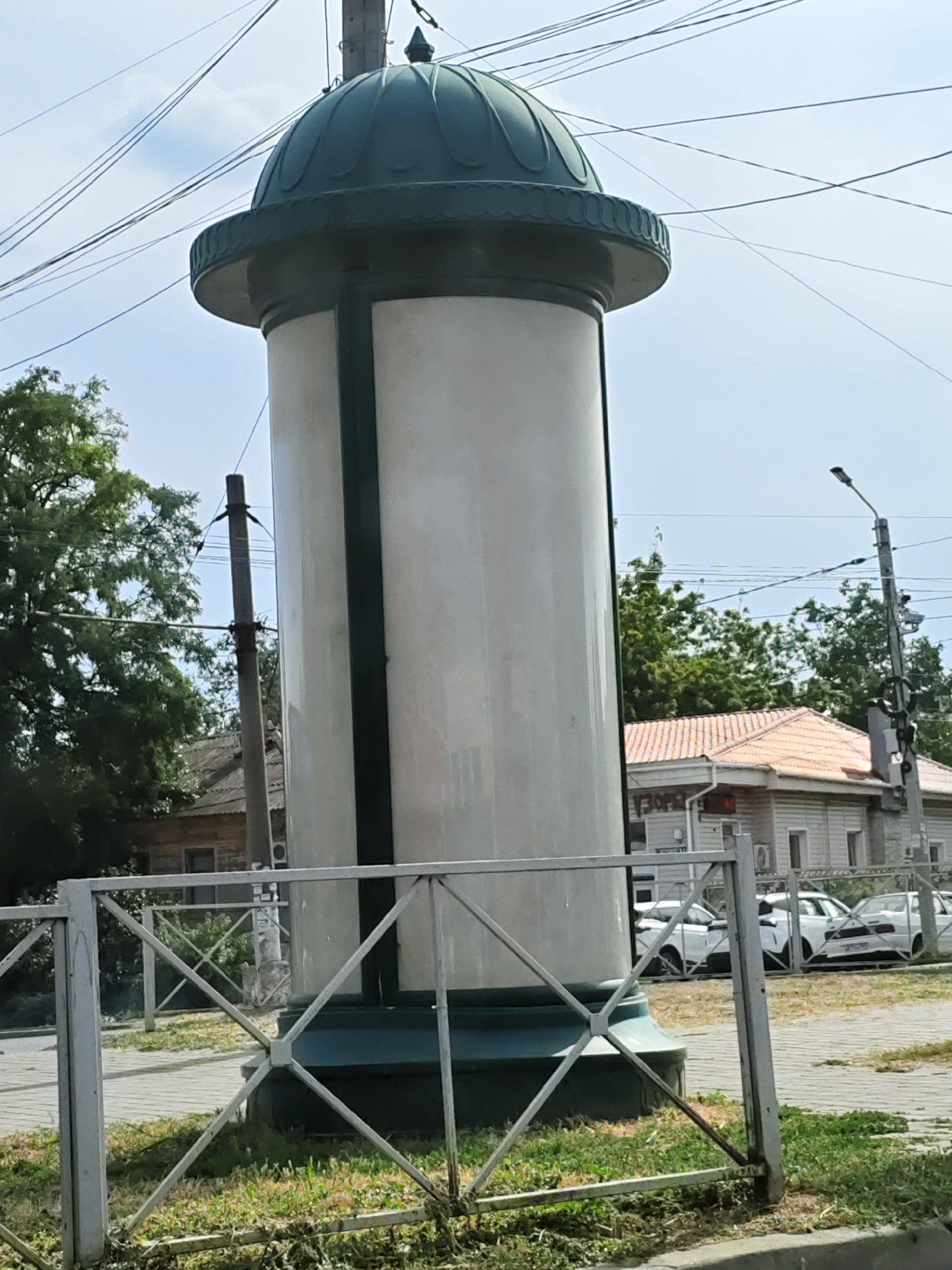 A tall, cylindrical billboard with a green domed top stands in the foreground, partially obscured by a decorative metal fence. The billboard's cylindrical body is white with a dark green vertical stripe. Behind the billboard, a street light on a pole with multiple wires crisscrossing the sky is visible. Further in the background, several white cars are parked on a paved area. A building with a light-colored tiled roof and light-colored walls is situated behind the cars. Trees with lush green foliage surround the scene. The overall environment suggests an outdoor urban setting, possibly a street or public square, in Novocherkassk, Russia.