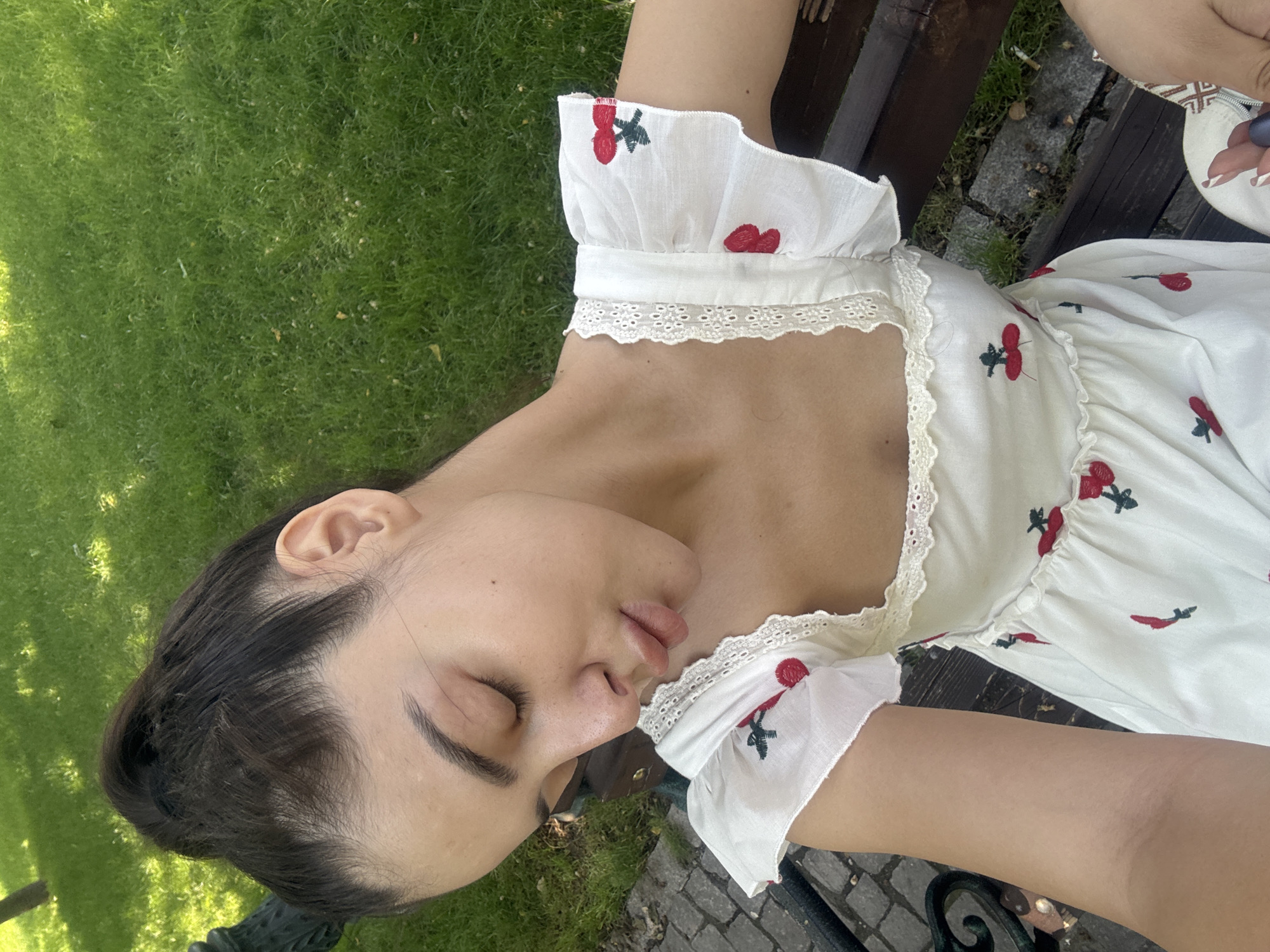 An outdoor, high-angle shot captures a young woman resting on a dark wooden park bench during the daytime. Her head is tilted to the side, with her eyes closed and lips slightly parted, suggesting she is asleep or deeply relaxed. Her dark hair is pulled back, revealing her ear.
She is wearing a white dress adorned with red cherry embroidery and delicate white lace trim along the neckline and puffed sleeves. The dress has a ruffled texture. Around her, vibrant green grass is visible, with patches of sunlight and shadow. Adjacent to the bench, there's a section of irregular cobblestone paving.
Partial views show her arm and shoulder. On the bench next to her, a small, light-colored pouch or bag with an embroidered pattern is visible, along with a dark purple, round object. Notably, a discarded cigarette butt lies on the cobblestone ground near the bench. The bright, natural lighting indicates it is sunny daylight, contributing to a peaceful and serene atmosphere. There is no visible text in the image.