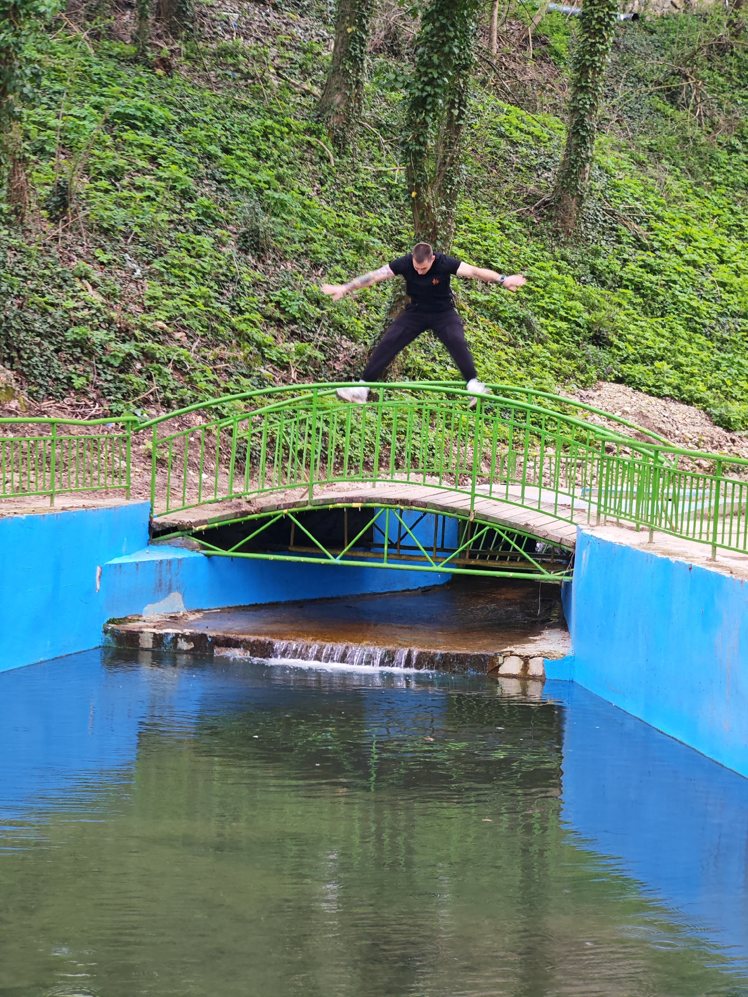 A man is jumping over a green bridge that crosses a blue canal. The bridge is made of wood and metal, and the canal has a small waterfall. The man is wearing black clothes and white shoes, and his arms are outstretched. In the background is a green hillside with trees. The location is Unknown, Unknown.