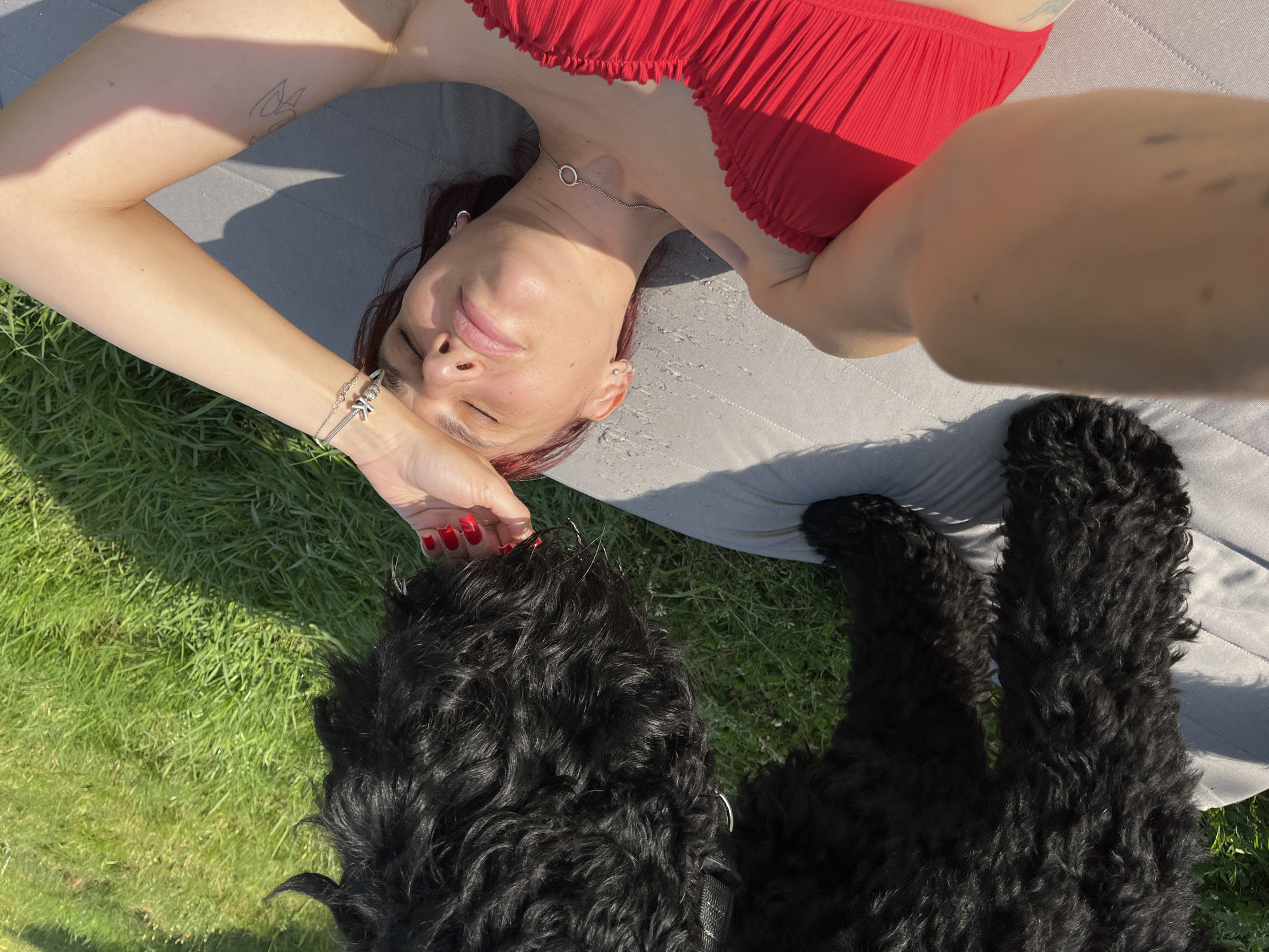 A person is lying on a gray mat outdoors, with a black dog standing over them. The person is wearing a red bikini top and has their eyes closed, with one hand on their forehead. The dog appears to be sniffing the person's hand. The ground is covered in green grass, and the scene is illuminated by sunlight.