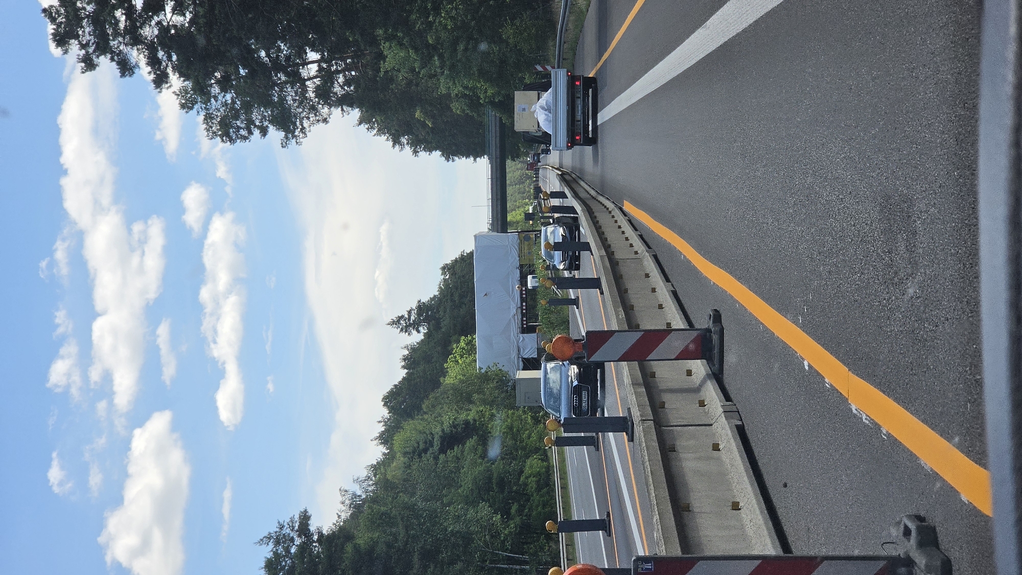 The image shows a view from a vehicle on a German highway, likely near Calau, Germany, given the context. The scene depicts a sunny day with scattered white clouds in a bright blue sky. The highway has multiple lanes, marked with white and orange lines. Construction barriers and orange warning lights are visible along the side of the road, indicating road work is in progress.
Several vehicles are present on the highway, including a white truck with a load covered in white plastic, and several passenger cars, including a dark colored Audi. The surrounding area is heavily wooded with tall green trees. A bridge or overpass is visible in the distance. The time of day appears to be daytime, possibly mid-morning or afternoon, due to the bright sunlight and clear skies. No people are clearly visible, but the presence of vehicles suggests traffic. The image captures a typical highway scene with a focus on the road and surrounding environment during what seems to be a period of construction.
