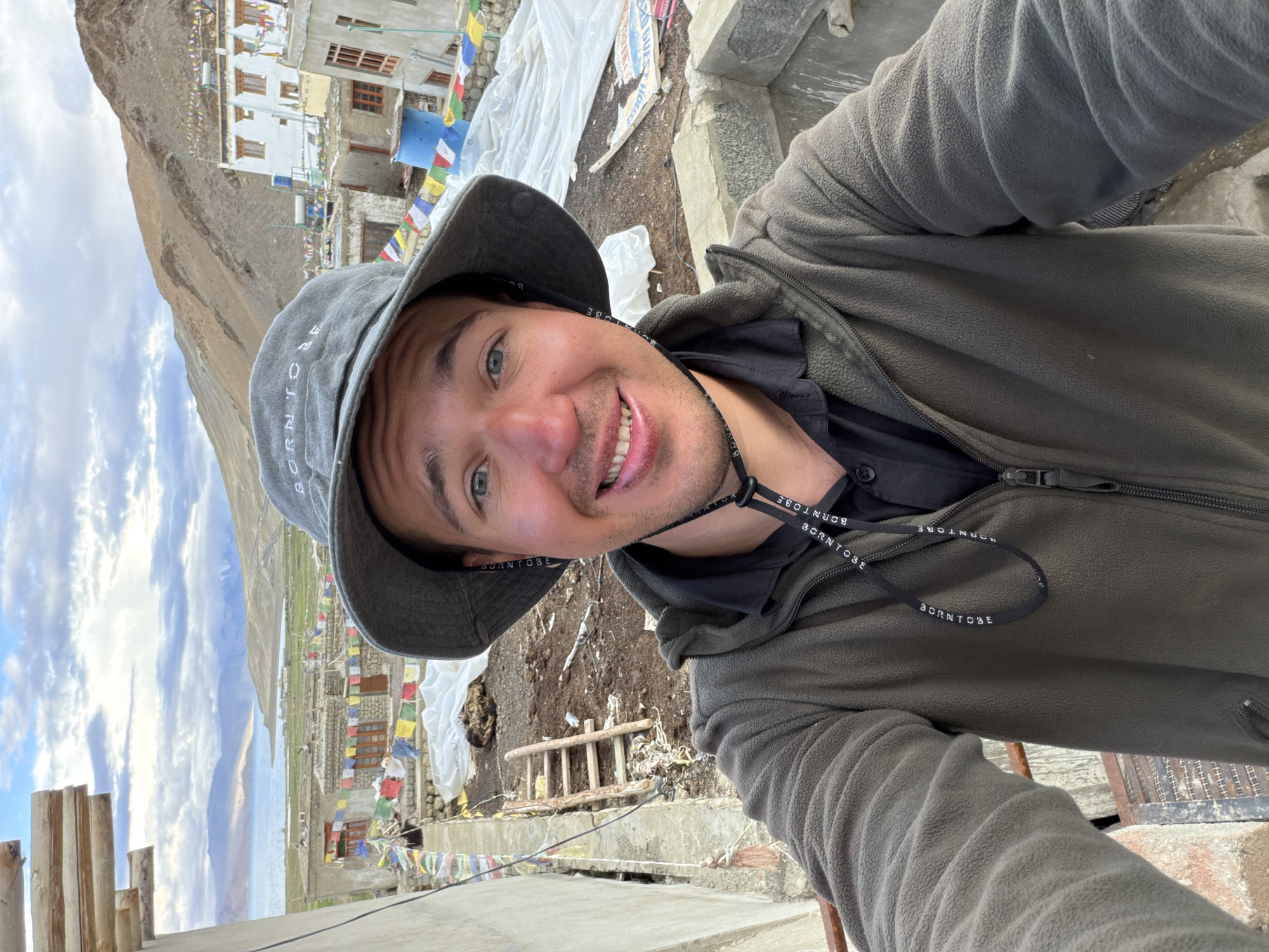 A man wearing a grey bucket hat and a dark green fleece jacket stands outside in what appears to be a village. The hat has the words "BORN TO BE" embroidered on it. He is looking at the camera and smiling. In the background, there are buildings decorated with prayer flags, a body of water, and mountains. The location is identified as Karzok, India. The scene suggests a tourist or traveler documenting their experience in a remote, mountainous region.