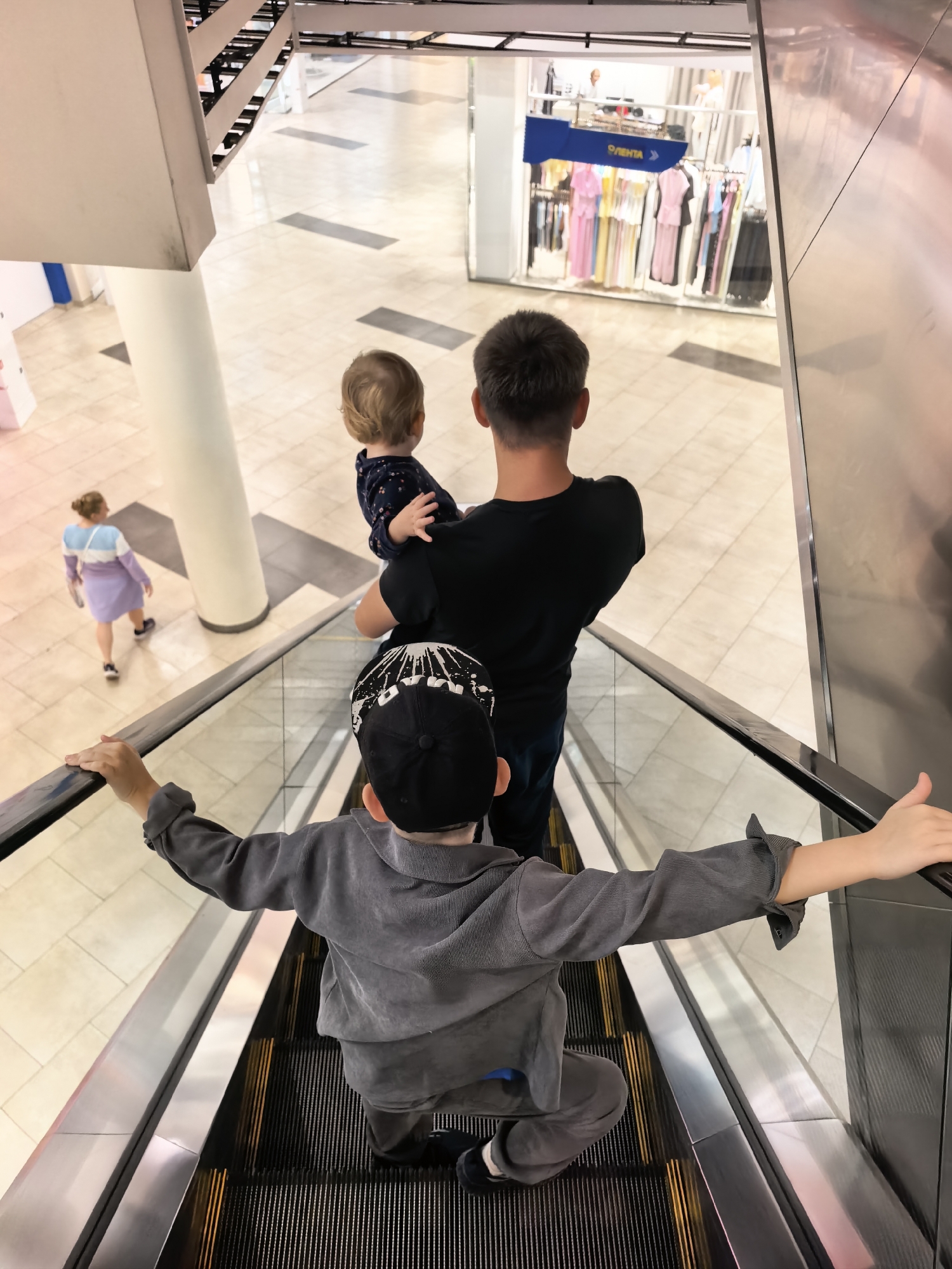 A family is on an escalator in a shopping mall. A father is carrying a baby on his shoulders, and an older son is in front of him. The older son is wearing a black baseball cap and a gray tracksuit. The father is wearing a black t-shirt. The baby is wearing a dark blue shirt with white polka dots. In the background, a woman wearing a light purple dress is walking away from the escalator. The mall has a polished tiled floor and clothing racks are visible in a store in the distance. The escalator has silver-colored metal railings and black treads with yellow warning stripes. The overall scene is brightly lit. The image was taken in the shopping mall "Otentia" in Sterlitamak, Russia.
