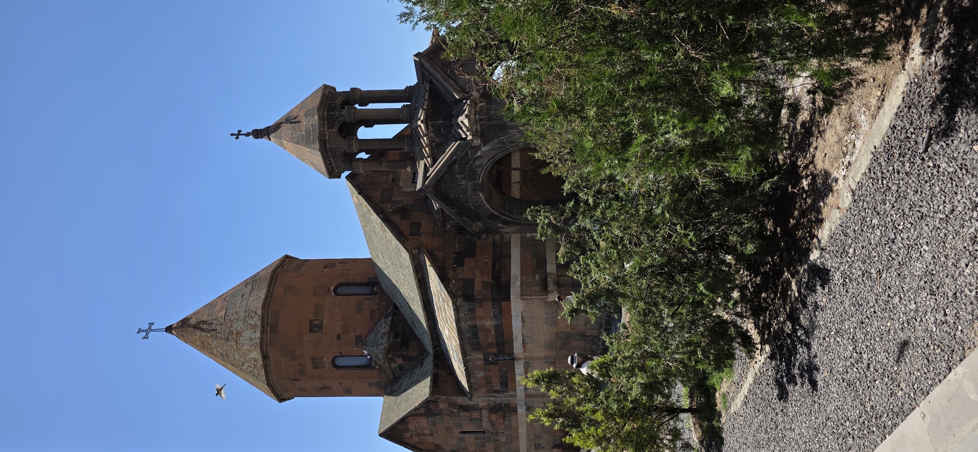 In Lusarat, Armenia, on a clear, bright day with a vibrant blue sky, an ancient church stands with its characteristic conical roof and bell tower. The church is constructed of reddish-brown stone, accented with darker bands, and features arched windows and a prominent cross atop its main dome. To the right, a smaller bell tower with an octagonal roof and open arches is visible. In the foreground, lush green trees and shrubs partially obscure the view of the stone pathway leading to the church. Two individuals, a person wearing a hat and another partially visible figure, can be seen near the church entrance, engaged in what appears to be a leisurely stroll. A bird is captured mid-flight in the upper left quadrant of the image, adding a dynamic element to the serene scene. The ground in the foreground is covered with dark gray gravel.