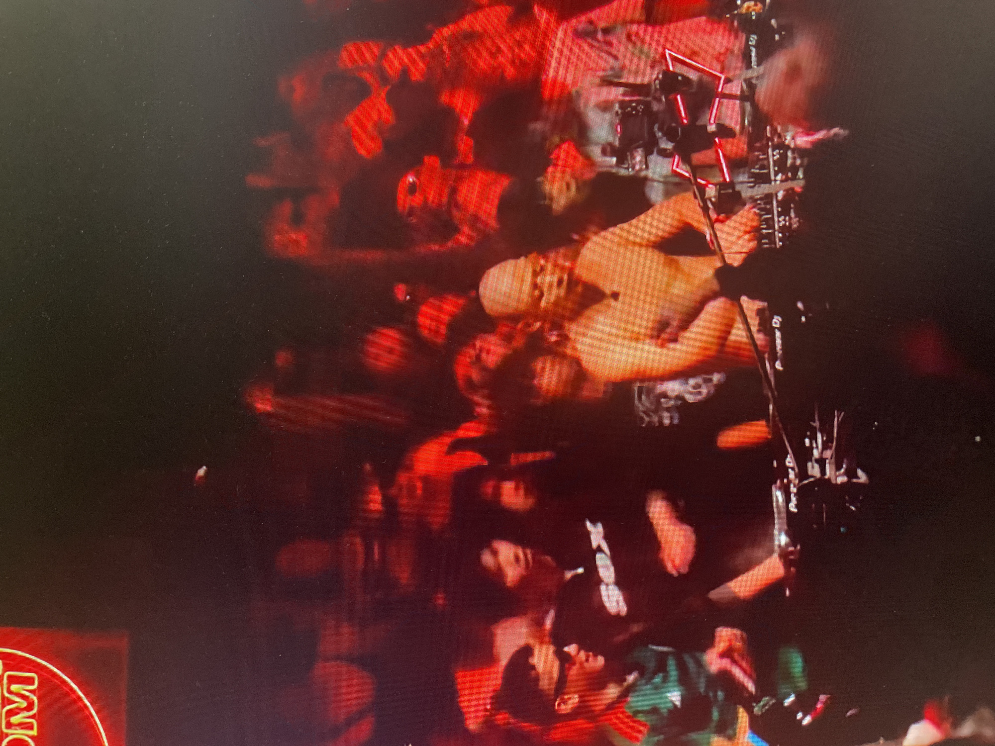 An outdoor, dimly lit festival scene in an unknown city in Russia shows a DJ at work. A bald, shirtless man, likely the DJ, is visible in the foreground, with his hands on a DJ console. He is facing a crowd of people, some of whom are illuminated by red lighting. The crowd appears to be enjoying the music, with some individuals raising their hands in the air. The DJ console has a "Pioneer DJ" logo visible on it. There is a neon sign in the background, partially obscured, with red lettering that appears to read "COM". The overall atmosphere suggests a lively music event.