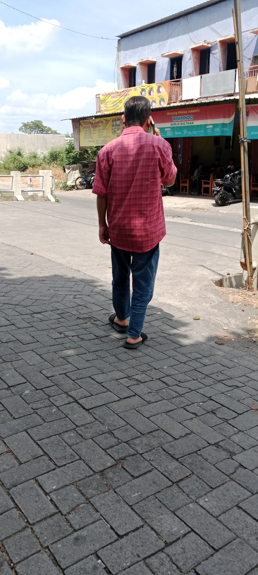 A person, viewed from the back, is walking on a gray brick sidewalk in front of a building. The person is wearing a red plaid short-sleeved shirt, blue jeans, and black flip-flops. The building appears to be a commercial establishment with multiple windows and awnings. A banner above an entrance reads "Warung Makan Indomie WARMINDO BURJO SULTAN". A motorcycle is parked on the street to the right of the building. The street is paved and there is a gray concrete barrier on the left side of the sidewalk. Trees and vegetation are visible in the background. The sky is blue with scattered clouds. The scene is likely located in Maguwoharjo, Indonesia, a suburban area known for its street food stalls and local businesses.