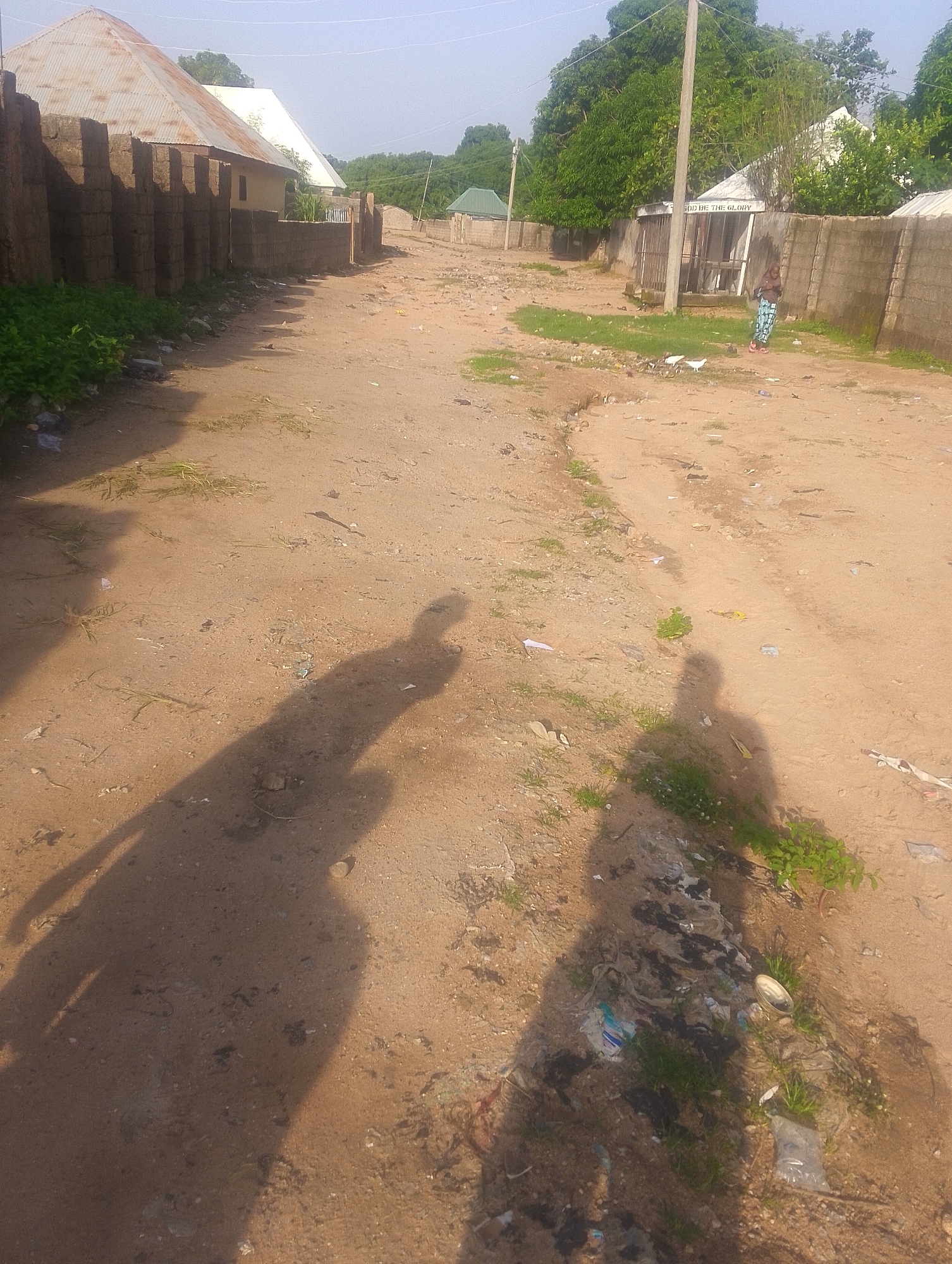 The image depicts a dusty, unpaved street in Jalingo, Nigeria, on a clear, sunny day. The scene is characterized by a worn dirt road winding through a residential area with modest buildings and a backdrop of lush green trees.
On the left side of the road, a fence made of stacked bricks borders properties with corrugated metal and white roofs. To the right, a similar brick fence runs along the street. In the mid-ground, a small building with a sign is visible; the text "GOD BE THE GLORY" is clearly legible on it. A person in a blue and white patterned garment is standing near this building, looking towards the ground. A few white birds are also present on the street.
The foreground of the image is dominated by the long shadows of two individuals, suggesting it is either early morning or late afternoon. The ground is sandy and shows signs of scattered litter, including plastic bags and other debris, mixed with patches of green grass and dry vegetation. The overall impression is a glimpse into the everyday life of a community in Nigeria, with a focus on the rustic setting and the presence of its inhabitants and their environment.