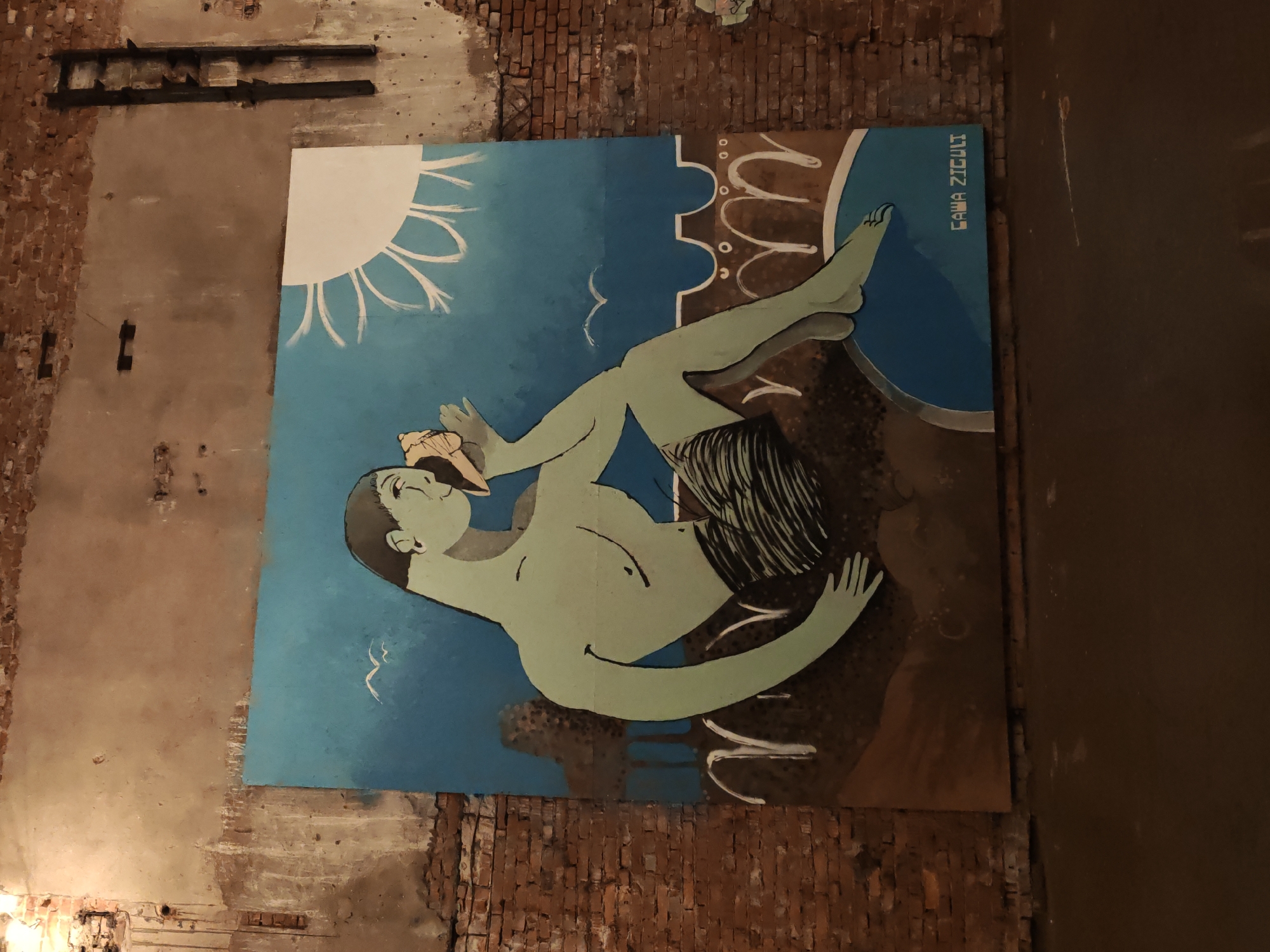 This is a photograph of a large mural painted on a brick wall in Saint Petersburg, Russia. The mural depicts a stylized, pale green figure with dark hair holding a seashell to its ear. The figure is seated on a brown, textured surface with a blue pool or water area in the foreground. In the background, there is a simplified depiction of a sun with radiating lines and some abstract forms. The mural is signed "GAWA ZICULI" in white lettering at the bottom right. The wall itself is made of weathered red brick with patches of crumbling plaster, suggesting an urban or industrial setting. The lighting in the image is dim, with some artificial light illuminating the mural from above.