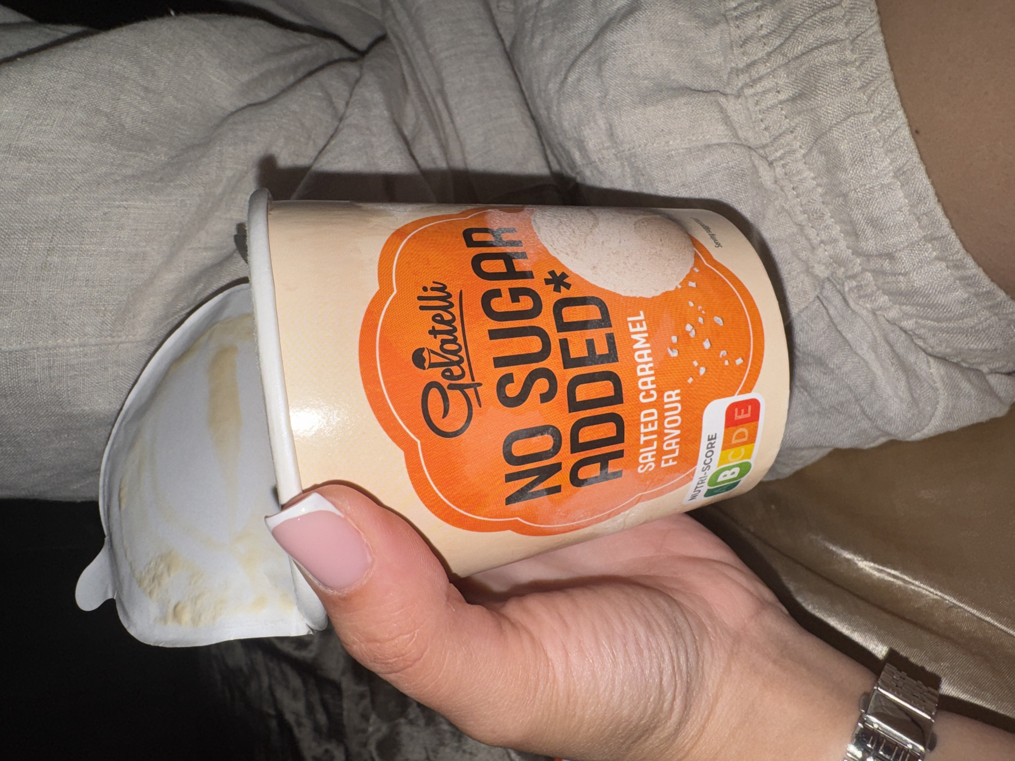 A close-up shot shows a person holding an open container of "Gelatelli NO SUGAR ADDED" salted caramel flavored ice cream. The container is partially open, revealing some of the ice cream inside and smears on the lid. The person's hand, with manicured nails, is holding the container. The background is out of focus, showing a neutral-colored fabric, possibly clothing or bedding. The image does not contain any elements that would allow for the determination of its location in Plovdiv, Bulgaria, or any specific city.