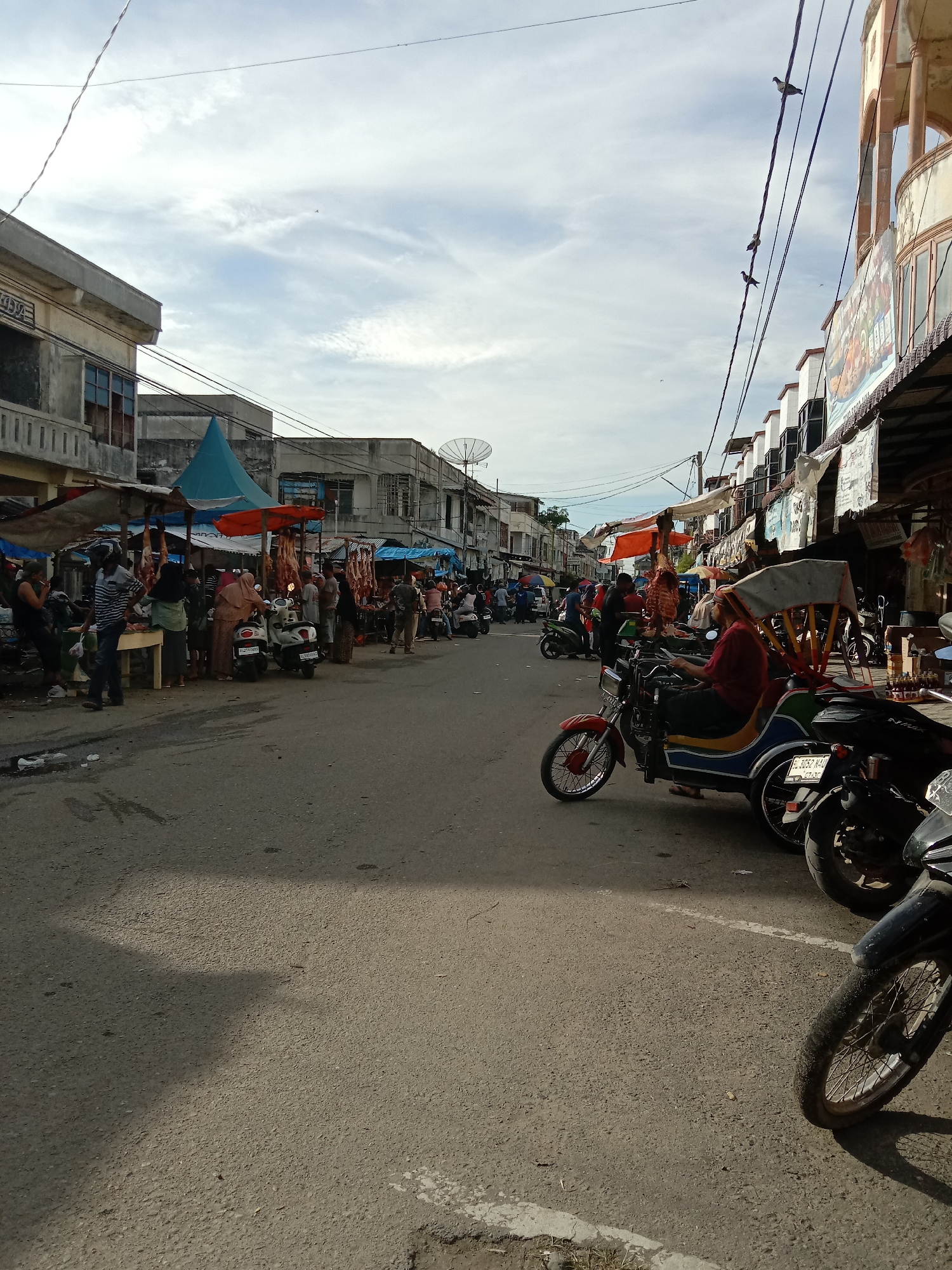 This image captures a street scene in Lhokseumawe, Indonesia. The scene depicts a bustling street market. People are milling about, some standing at stalls where meat is hanging. A number of motorcycles are parked on the right side of the street. A three-wheeled vehicle is visible, with a driver sitting inside. Buildings line both sides of the street, some with awnings. Overhead, wires crisscross the sky. The sky is partly cloudy.