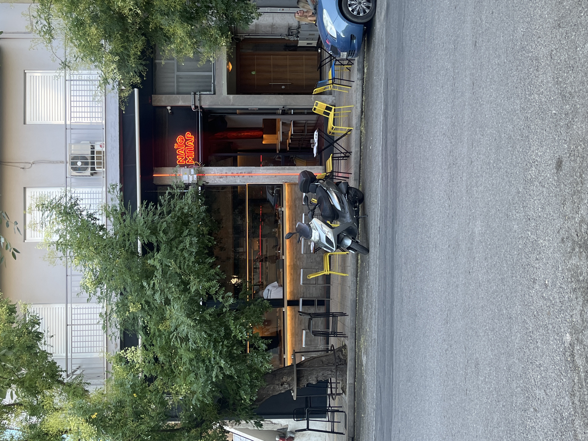 In Athens, Greece, a street scene unfolds with a focus on a cafe with a prominent neon sign that reads "NAIO MΠAP". The cafe features an inviting outdoor seating area. A grey scooter is parked in front of the cafe, and to the right, a blue car is visible. On the cafe's balcony, an air conditioning unit is mounted on the wall. Green trees provide shade and natural beauty to the urban environment.