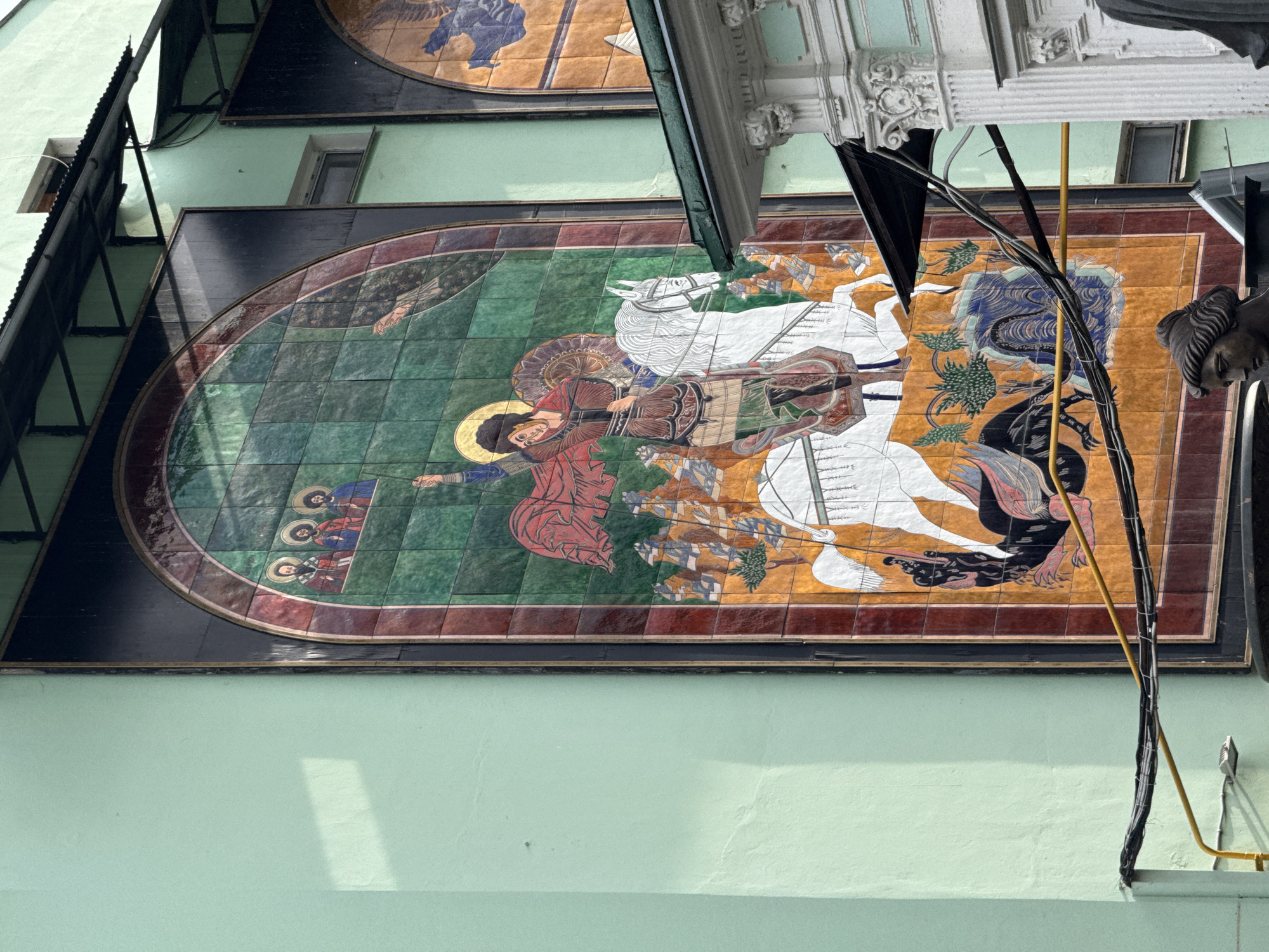 This image shows a large, oval mosaic mural on the side of a pale green building. The mural depicts a religious scene, likely from Orthodox Christian iconography. The central figure is a warrior saint, possibly Saint George or Saint Demetrius, on horseback, wearing a red cloak and a golden halo. He appears to be fighting a dragon or a dark, monstrous figure. Another horse, ridden by a figure in white, is also present, suggesting a narrative of triumph over evil.
To the left of the central scene, a smaller group of figures with halos are depicted, possibly saints or religious leaders. The mural is set against a background of green, brown, and orange tiles, arranged in a mosaic pattern. The overall style is traditional and stylized, characteristic of Eastern Christian art.
The mural is mounted on an exterior wall, and the lighting suggests it is daytime. There are no visible weather cues, but the building's pale green paint and the presence of electrical wires and conduits suggest an urban setting, likely Moscow, Russia, as indicated by the provided context. The artwork is framed by dark borders and a reddish-brown tiled border. Architectural elements, including decorative white moldings and a dark overhang, are visible at the top of the image, suggesting the mural is part of a larger building facade. The image is taken from a low angle, looking upwards at the mural.