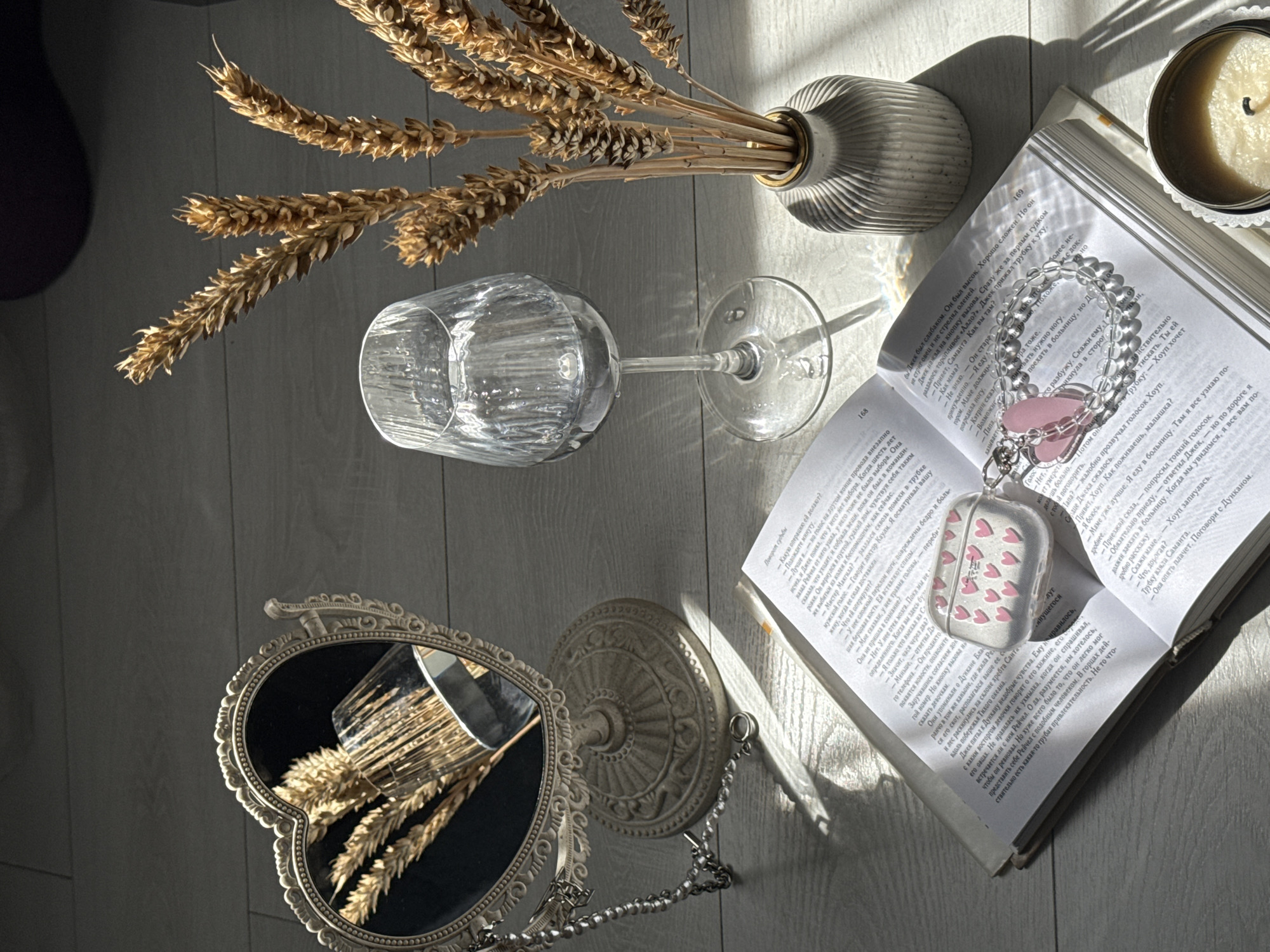 The image displays a still life arrangement on a light-colored wooden surface, likely a floor or table. A heart-shaped mirror with an ornate, antique-style frame sits on the left, reflecting a bouquet of dried wheat stalks in a glass. Adjacent to the mirror is a clear wine glass filled with water, refracting light. To the right of the glass, a grey ceramic vase holds a bundle of dried wheat stalks, similar to those in the mirror's reflection. In the foreground, an open book lies flat, its pages filled with text. Resting on the open book is a clear, heart-shaped case, possibly for earbuds, adorned with small pink hearts and a chain with a pink heart charm. A portion of a lit candle in a tin container is visible in the bottom right corner. The scene is bathed in natural light, creating strong shadows and highlights across the objects and surface. The overall ambiance suggests a quiet, possibly domestic setting. No people are present in the scene.