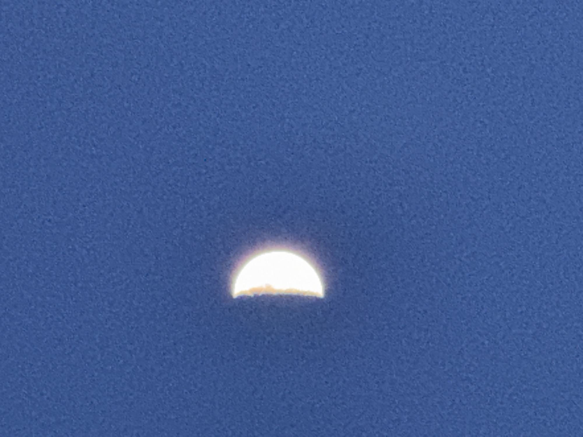 The image presents a striking view of a brightly illuminated half-moon against a clear, deep blue sky. The moon is centered in the lower half of the frame, glowing with an intense white light. Its lower, flat edge appears slightly indistinct and is touched with a faint brownish-orange hue, possibly indicating thin atmospheric haze, dust, or a wispy cloud layer partially obscuring its lower limb. The upper, curved portion of the moon is sharply defined. The surrounding sky is a uniform, vivid blue, suggesting that the photograph was taken during daylight hours or early twilight when the moon is still visible. The image exhibits a noticeable grainy texture throughout the sky, characteristic of digital noise or atmospheric conditions. No other celestial bodies, geographical features, or man-made objects are visible, and there is no discernible text. The scene depicts a calm and clear weather condition. While the location context is Chaikovsky, Russia, there are no specific visual cues within the image itself to confirm this.