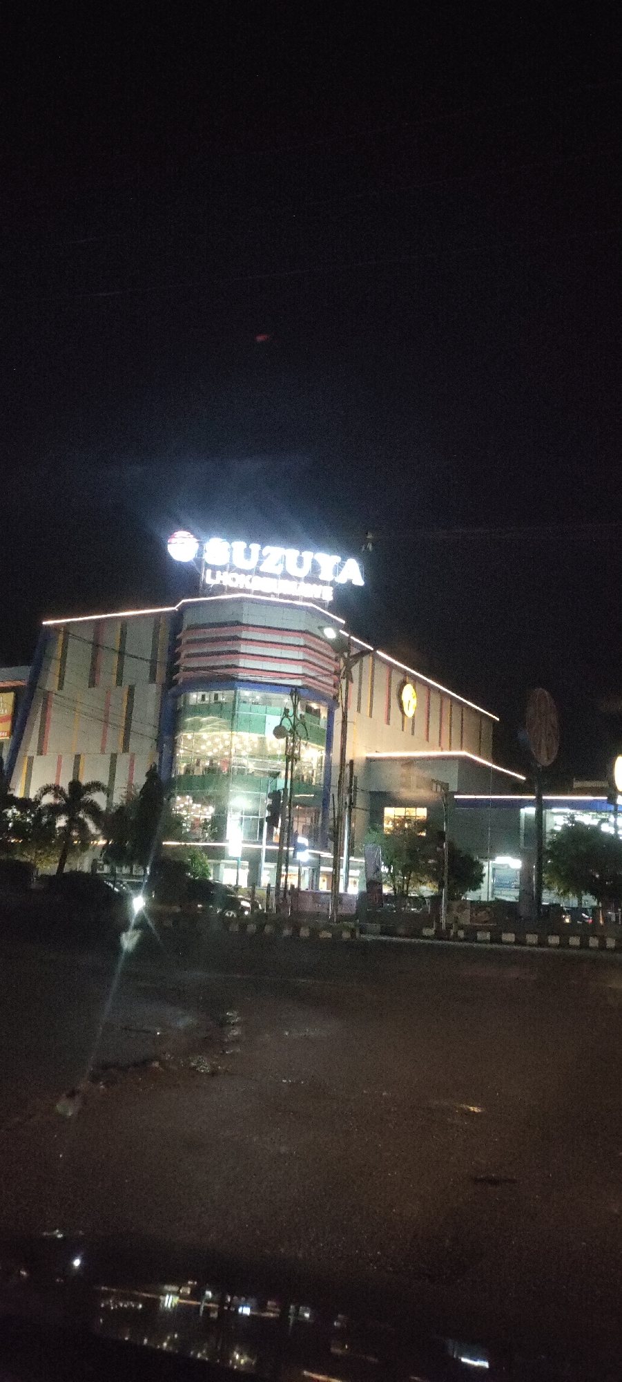 At night, a multi-story building labeled "SUZUYA Lhokseumawe" is visible, presumably located in Lhokseumawe, Indonesia. The building is brightly lit with both interior lights visible through glass windows and exterior decorative lighting along the edges. In front of the building is a street that appears wet, reflecting the surrounding lights. Several trees and streetlights are also visible in the foreground. The sky is dark, suggesting a late evening or night setting. A small, out-of-focus vehicle or light source is visible on the left side of the frame.