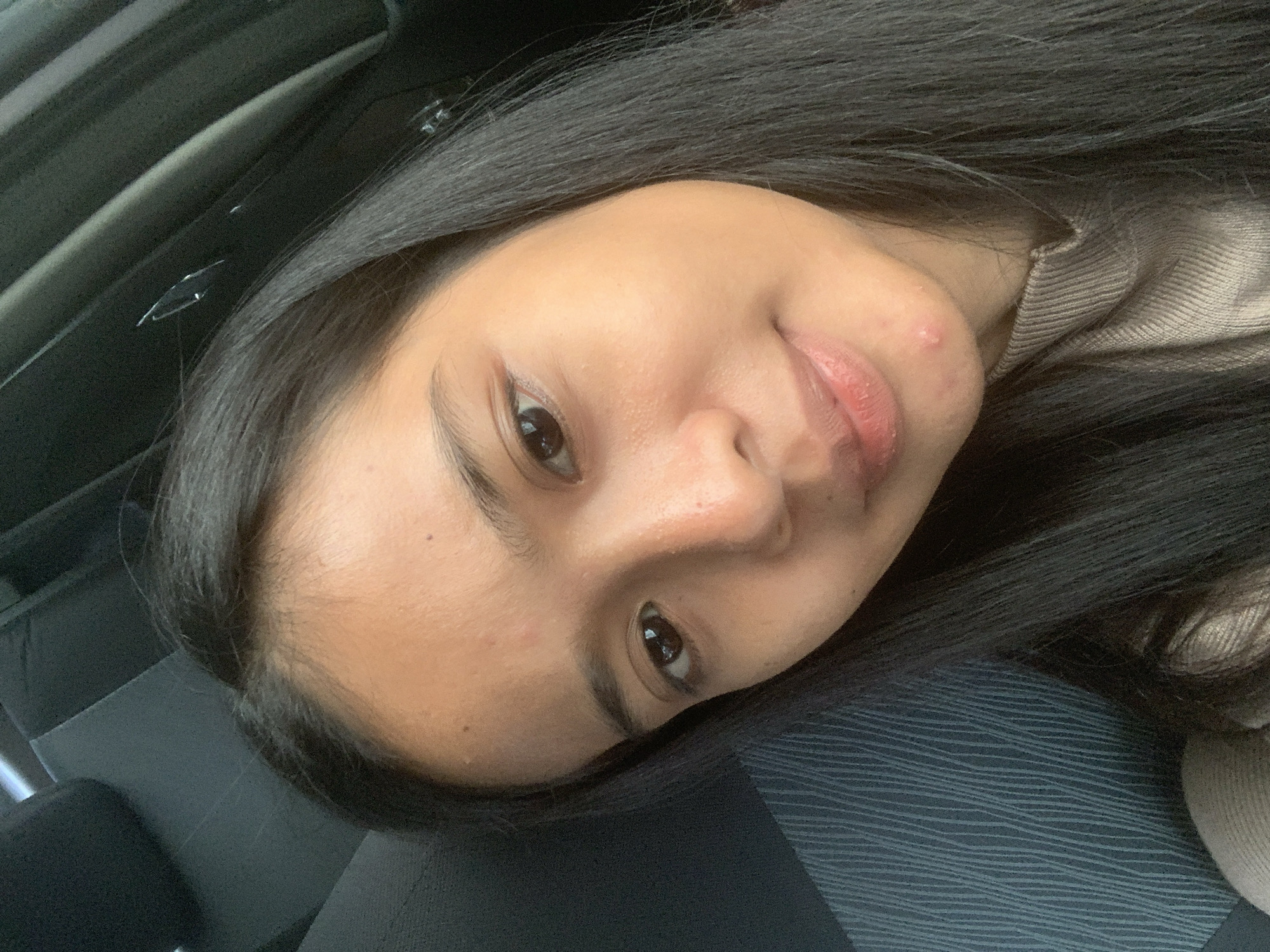 A close-up, slightly angled portrait shows a young woman with long, dark hair. She has brown eyes and fair skin. A noticeable blemish is present near the corner of her mouth. The background is indistinct, suggesting she is seated in a car, with the ceiling and textured seat visible. The location is unknown; the city of Samarinda, Indonesia, cannot be confirmed.